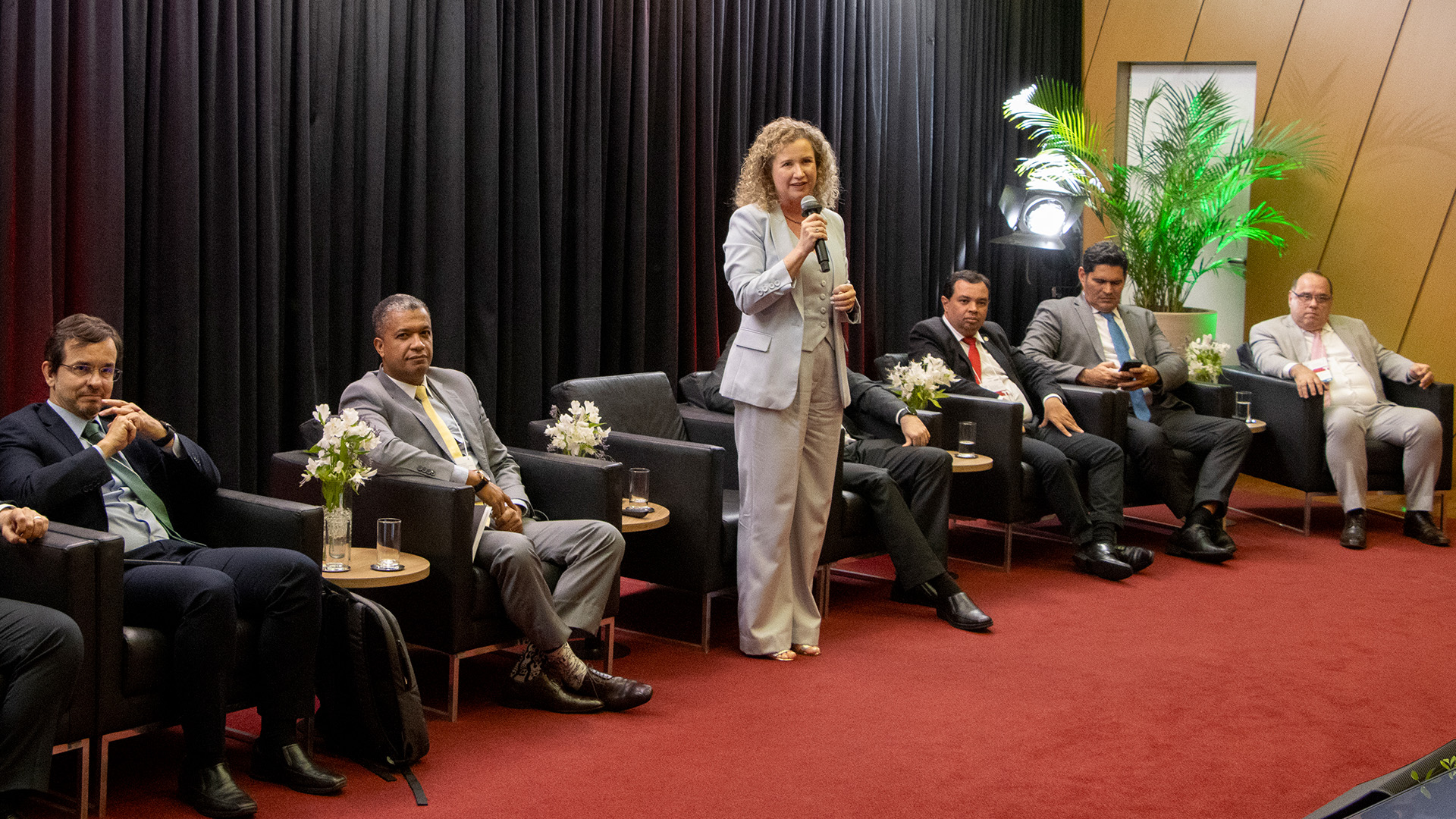 Foto da procuradora-geral do TCE/SC, Gláucia Mattjie, no palco do auditório do TCE/SC, falando ao público. Ela está em pé, ao centro. É uma mulher branca, de cabelos loiros e cacheados. Ao seu lado, demais integrantes que compõem o palco, apresentando as boas práticas.