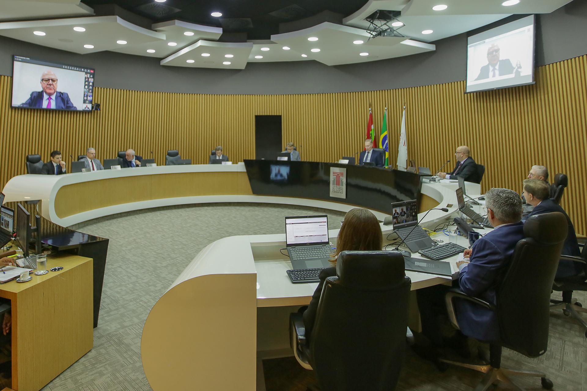 Foto horizontal da sessão do Pleno do TCE/SC. Aparecem os conselheiros que estão participando presencialmente. Na televisão, aparece o conselheiro Wilson Wan-Dall, que participou virtualmente.