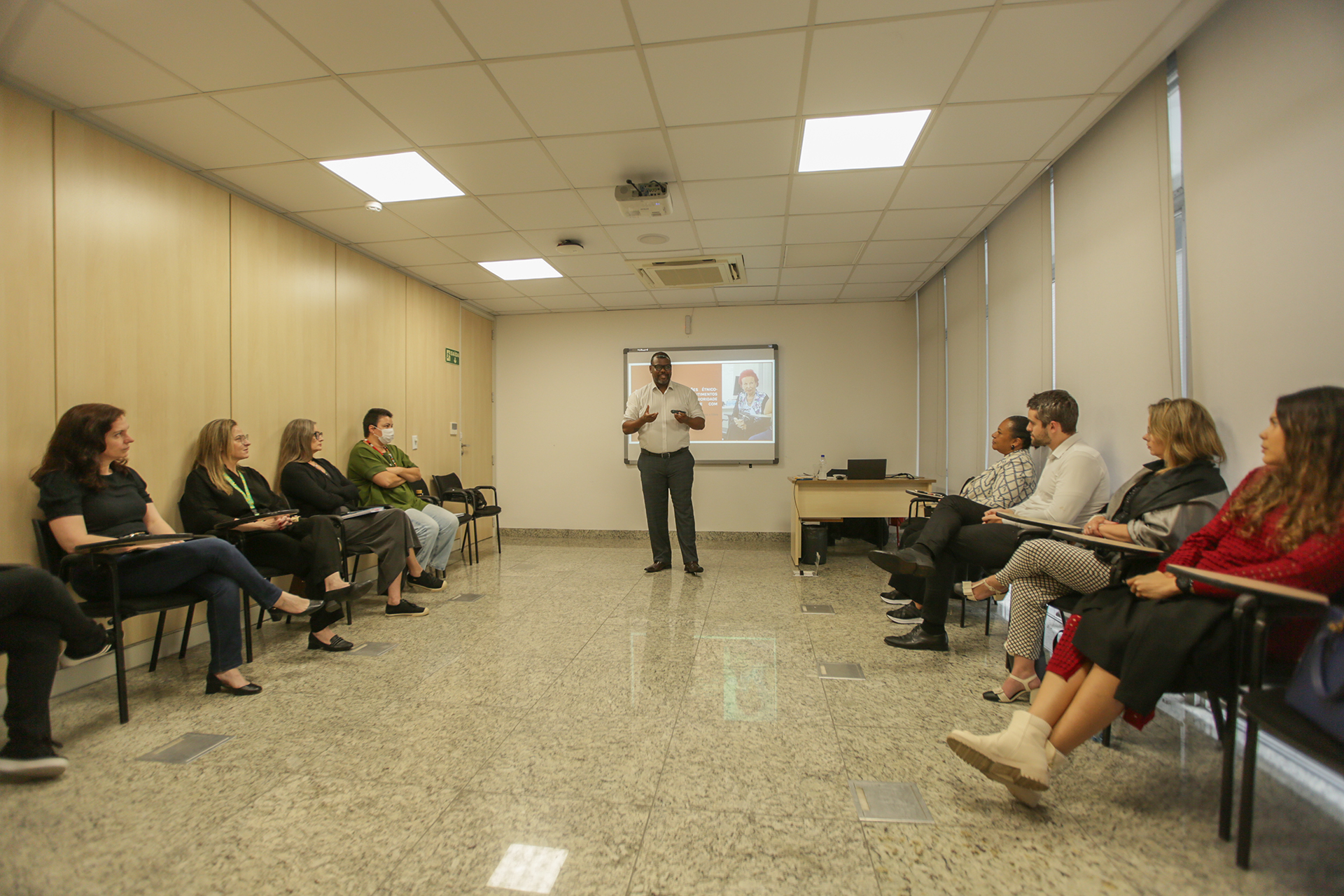 Foto que mostra, nas laterais, os participantes do terceiro módulo do curso, sentados em cadeiras pretas. Ao centro e ao fundo, o professor Delton Felipe, em pé, falando ao público. Atrás dele, é projetada uma apresentação. 