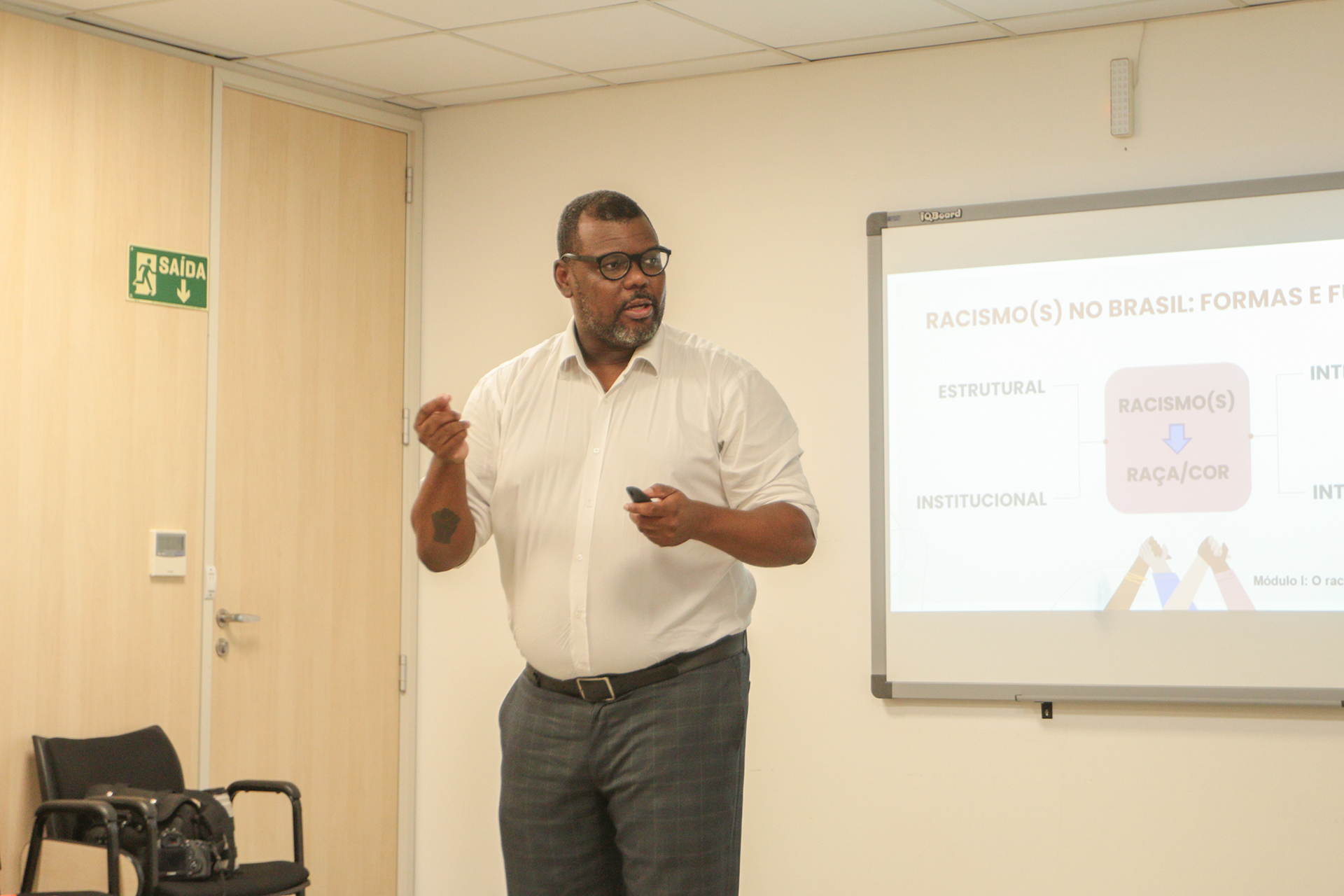 Foto do professor Delton Felipe ministrando o terceiro módulo. Ele é um homem negro, de cabelos e barba pretos. Veste camisa branca e está em pé. Atrás dele, projeção de apresentação. 