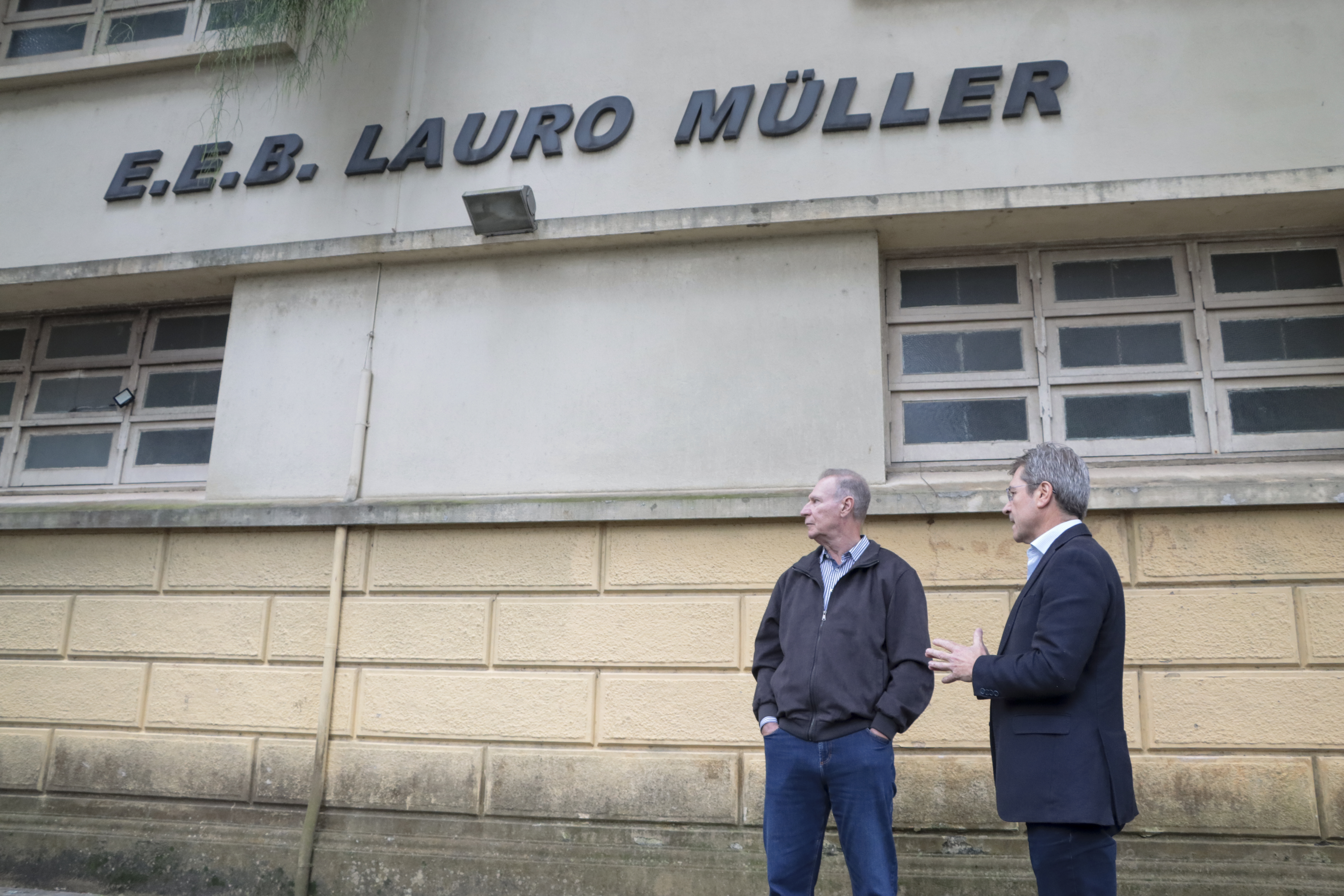 Foto horizontal do presidente e do vice-presidente do TCE/SC, conselheiros Herneus De Nadal e José Nei Ascari, respectivamente. Eles caminham pela parte externa do prédio da Escola Básica Lauro Muller.