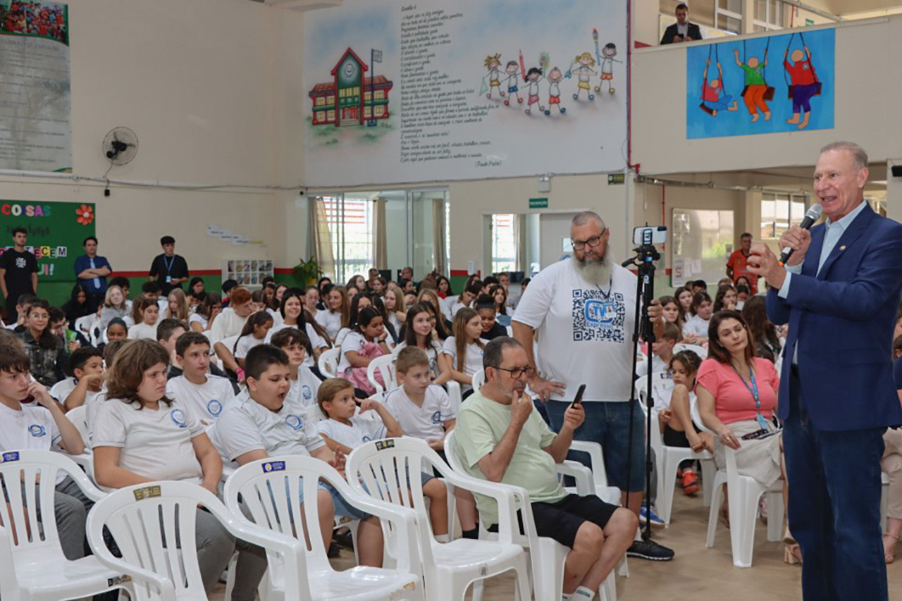 Foto horizontal em auditório escolar, com grande público sentado em cadeiras plásticas brancas. Ao fundo e nas laterais, há murais ilustrados com desenhos infantis e textos na parede. À direita da imagem, o presidente Herneus De Nadal aparece em pé, usando blazer azul, segurando um microfone e falando ao público. 