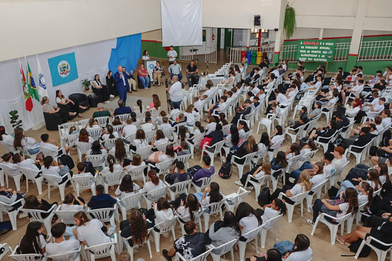 Foto horizontal aérea em auditório escolar, com grande público acomodado em cadeiras plásticas brancas organizadas em fileiras. À frente do espaço, há mesa com autoridades e área destinada às falas, com bandeiras e elementos institucionais ao fundo. Ao centro, o presidente Herneus De Nadal aparece em pé, usando blazer azul, dirigindo-se ao público. 