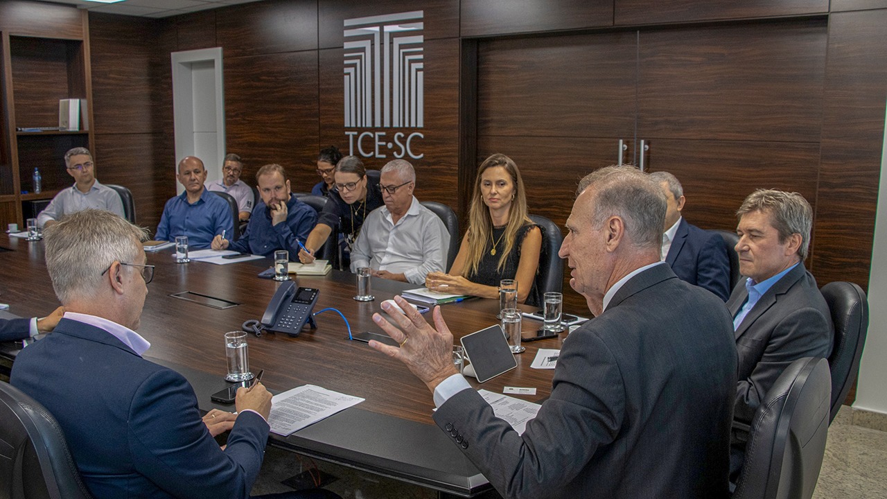 Banner horizontal com foto de reunião institucional entre integrantes do TCE/SC e representantes de agências reguladoras, realizada em sala de conferências com mesa retangular de madeira e o logotipo do TCE/SC ao fundo. Os participantes estão sentados ao redor da mesa, com documentos, copos de água e equipamentos de áudio distribuídos sobre a superfície. Em destaque, o presidente Herneus conduz a fala aos demais presentes. Também há a presença do conselheiro José Nei Ascari entre os participantes. 