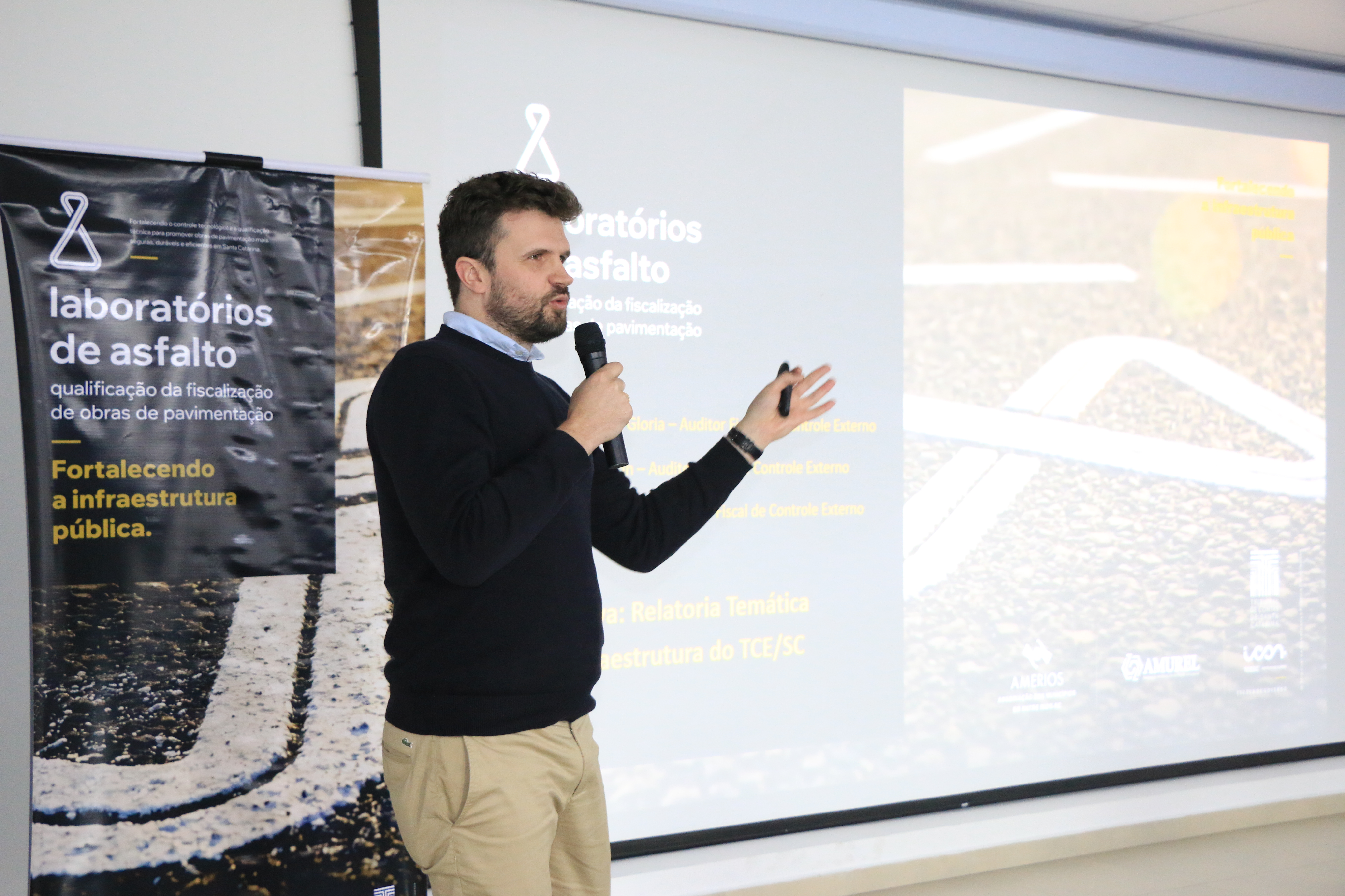 Foto horizontal do auditor Cléber Faccin falando ao microfone no evento. É um homem branco, de cabelos castanhos. Veste suéter preto. Ao fundo, slides são projetados com o tema "laboratórios de asfalto".