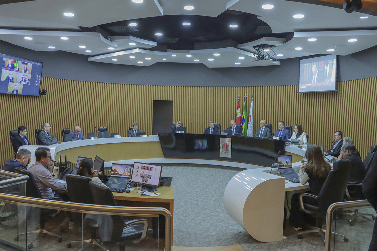 Foto ampla que mostra a composição da bancada circular durante sessão plenária. Ao centro, o presidente Herneus conduz os trabalhos. Nas laterais, os conselheiros e outras autoridades.