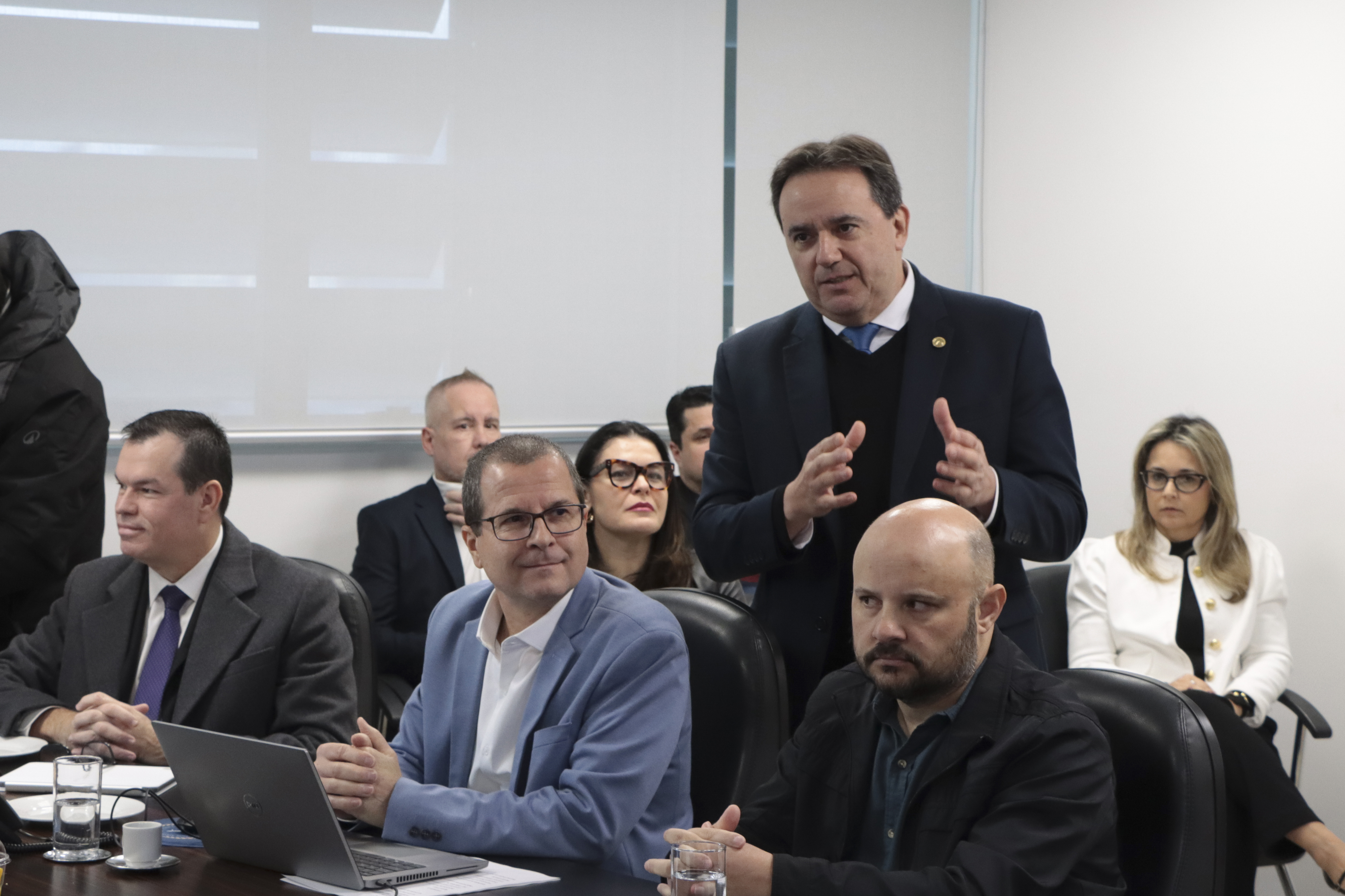 Foto horizontal do diretor de Comunicação Social do TCE/SC, João Cavallazzi, falando aos demais durante reunião. Ele está em pé, atrás de alguns integrantes sentados diante da mesa. Ao fundo, outros participantes. Entre os presentes, o conselheiro Adircélio e o diretor Nilsom Zanatto.