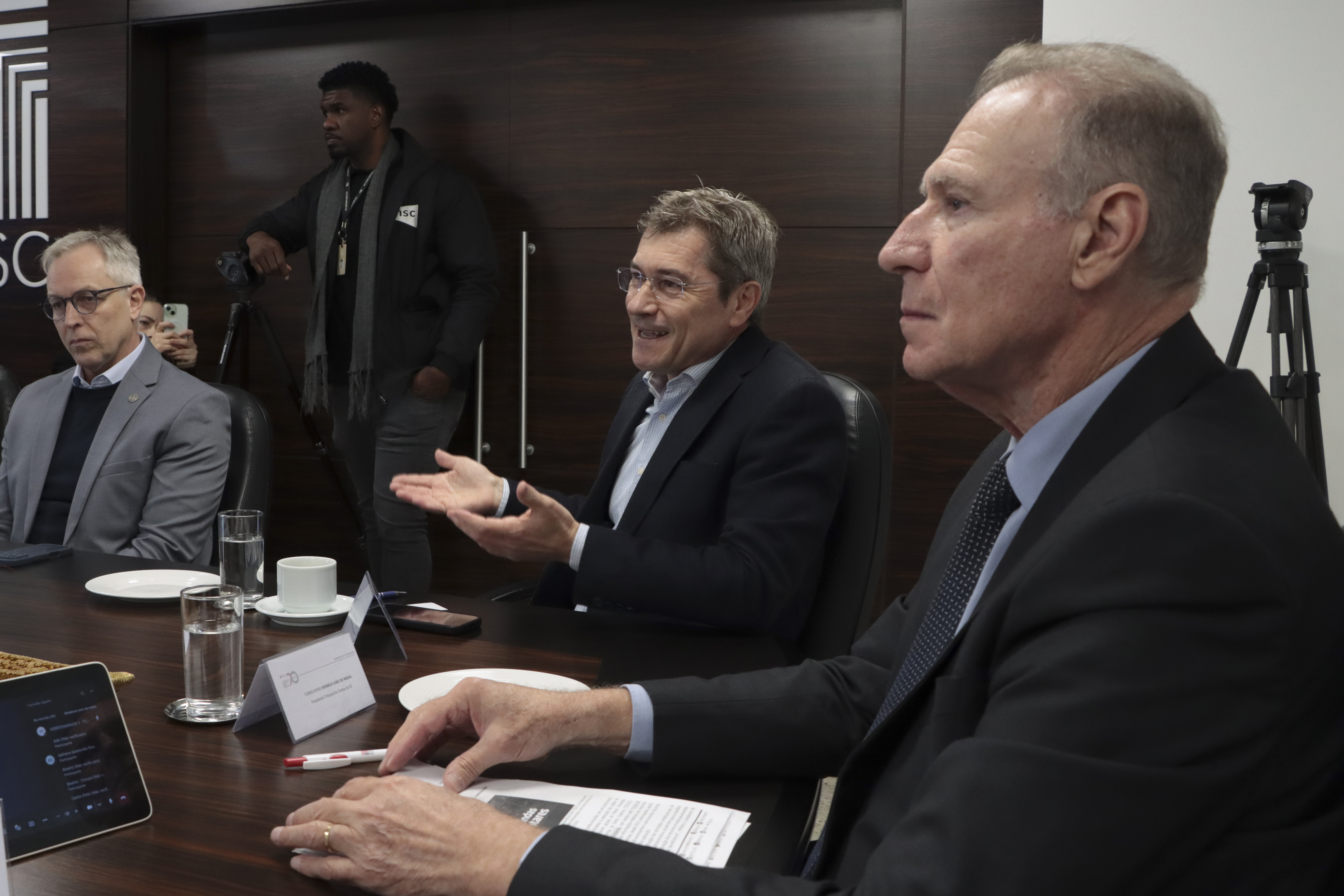 Foto horizontal do conselheiro José Nei Ascari, ao lado do presidente Herneus, falando aos demais durante reunião. Ao seu lado, também aparece o diretor Sidney Tavares Júnior. Eles estão sentados diante da mesa de reunião.
