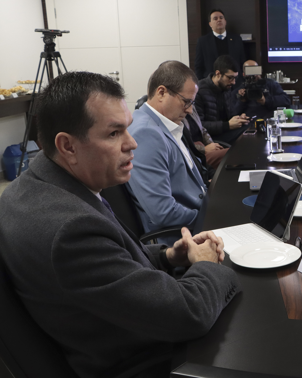 Foto vertical do corregedor-geral, conselheiro Adircélio de Moraes Ferreira Júnior, falando aos demais durante reunião. Ele está sentado diante da mesa, ao lado do diretor Nilsom Zanatto e de demais participantes.