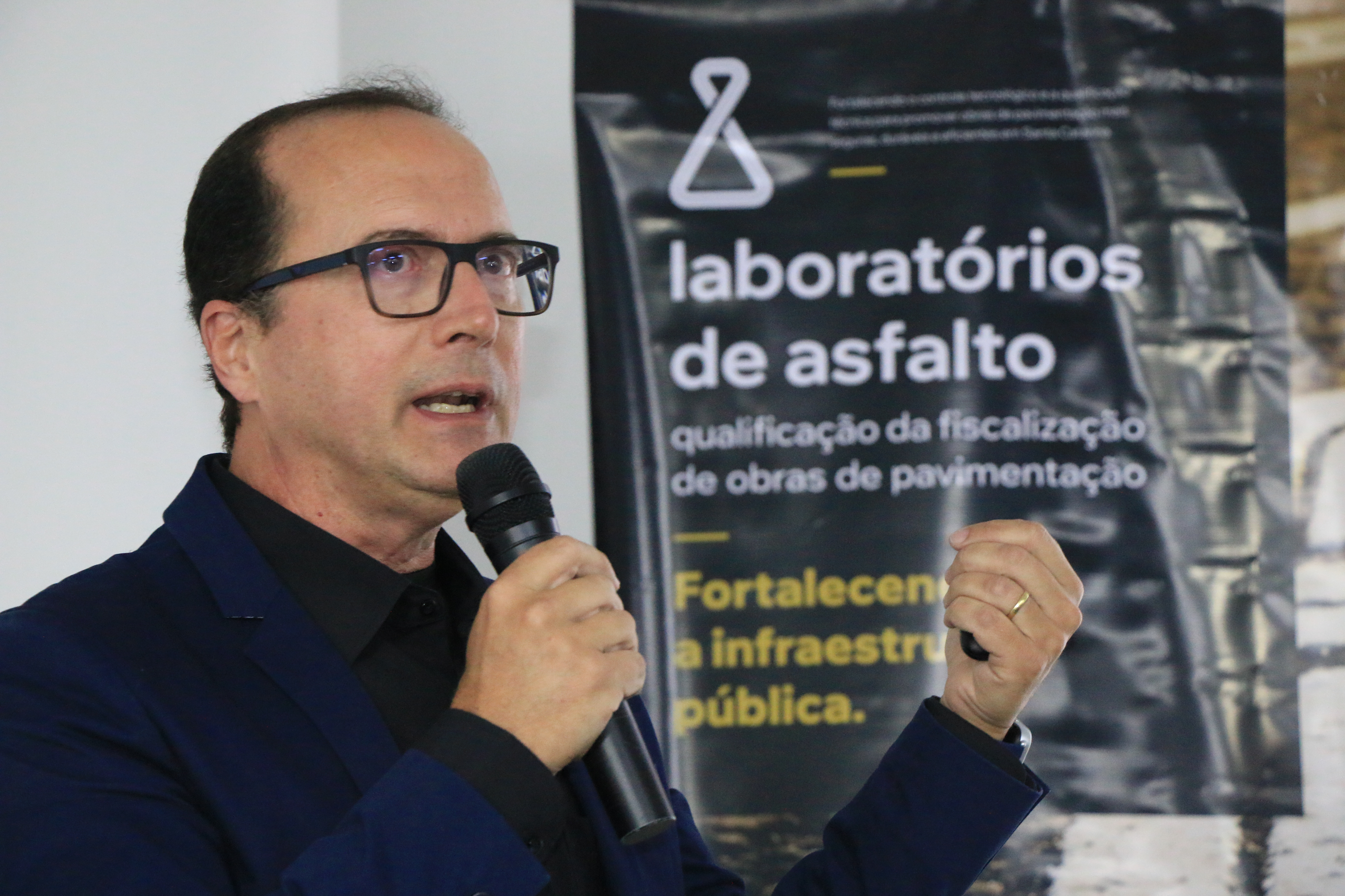 Foto horizontal do auditor Rodrigo Glória falando ao microfone no evento. É um homem branco, de cabelos pretos. Veste paletó azul-escuro e usa óculos de grau. Atrás dele, banner com logo do evento.