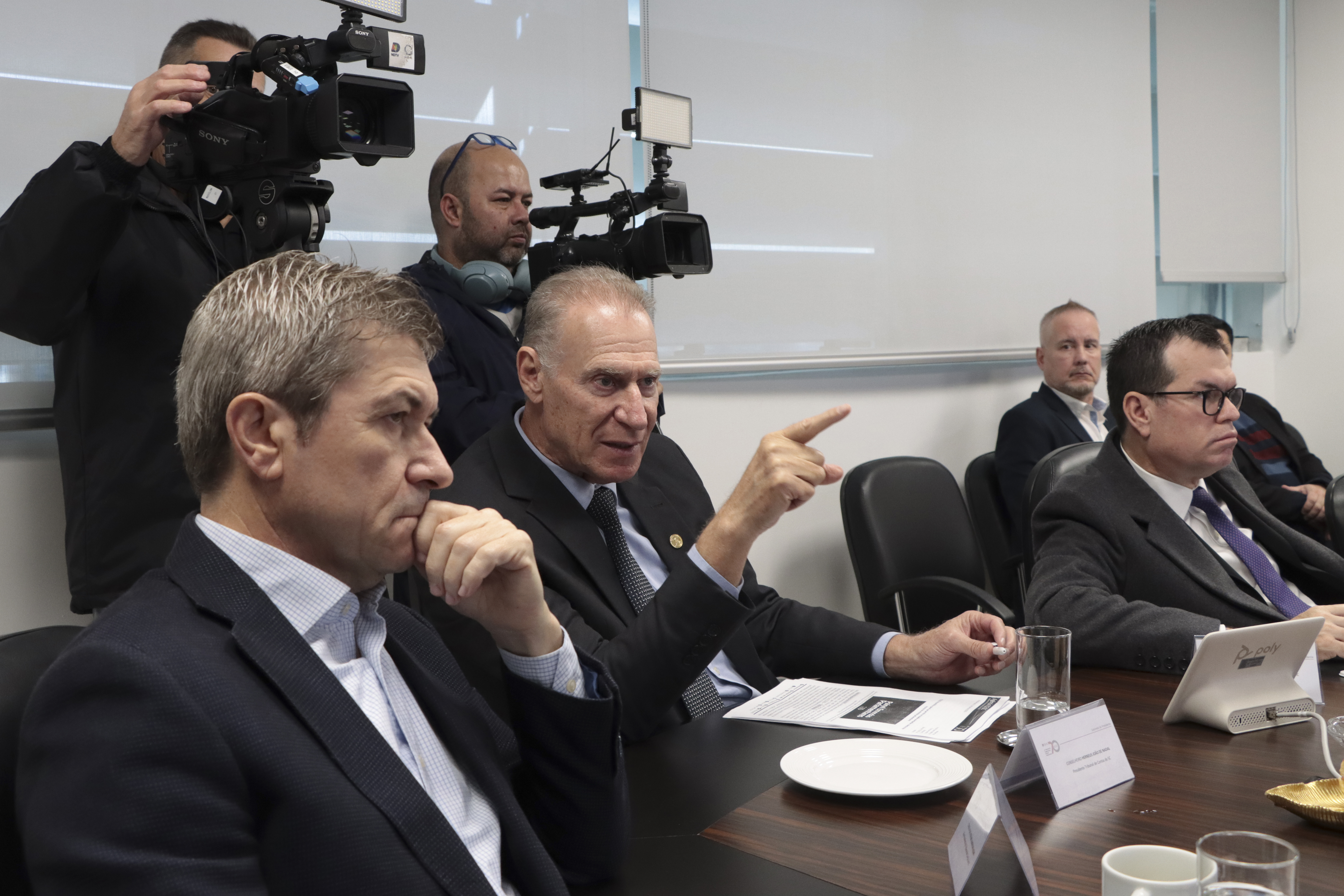 Foto horizontal que destaca o presidente do TCE/SC, conselheiro Herneus De Nadal, ao lado do vice-presidente José Nei Acari e do corregedor Adircélio de Moraes Ferreira Júnior. Eles estão sentados diante da mesa de reuniões. Herneus fala aos demais. Atrás, aparecem alguns operadores de câmera. Sobre a mesa, há papéis, placas de identificação e copos de água.