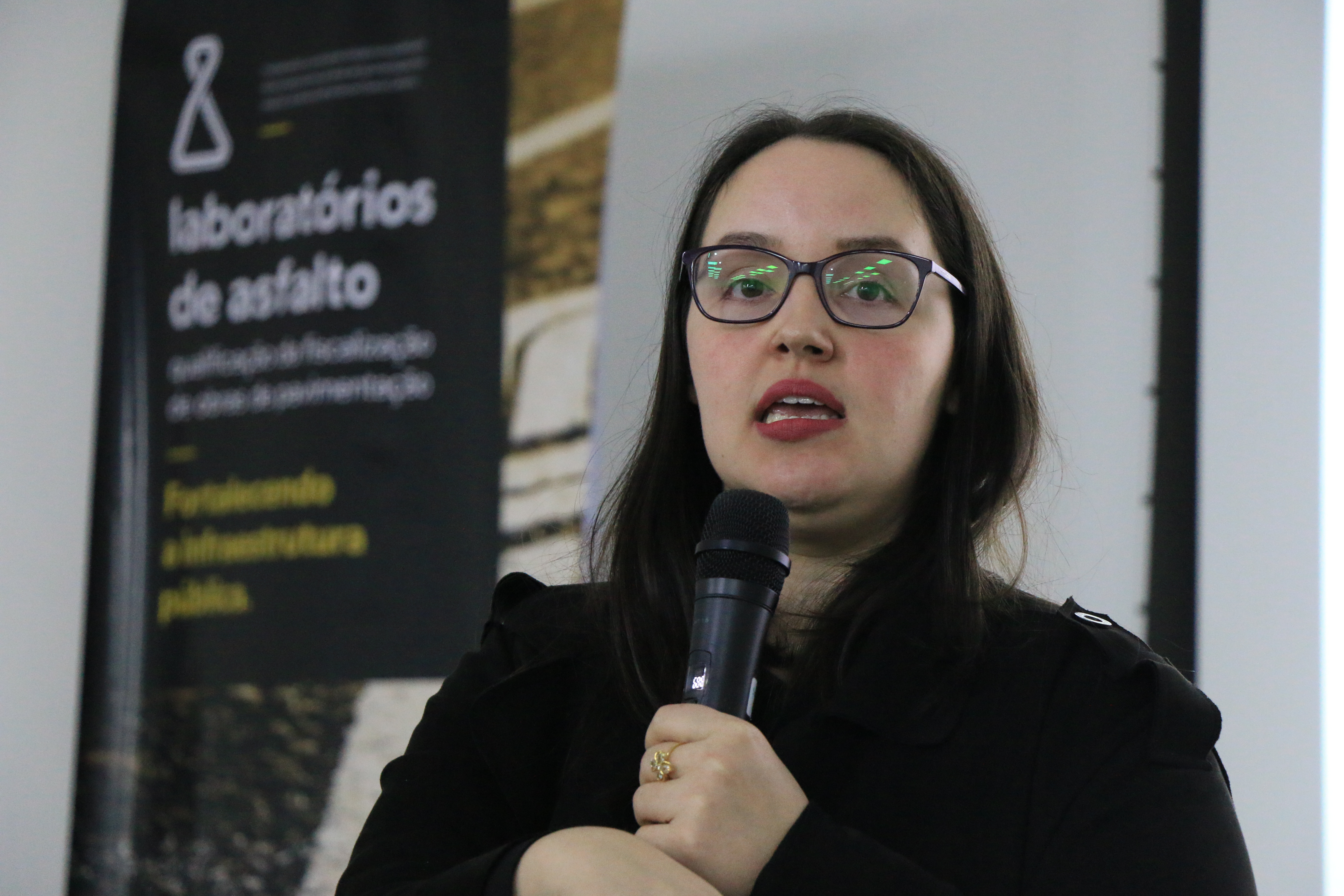 Foto horizontal da auditora Renata Ligocki Pedro. Ela está  falando. Atrás dela o banner do evento.