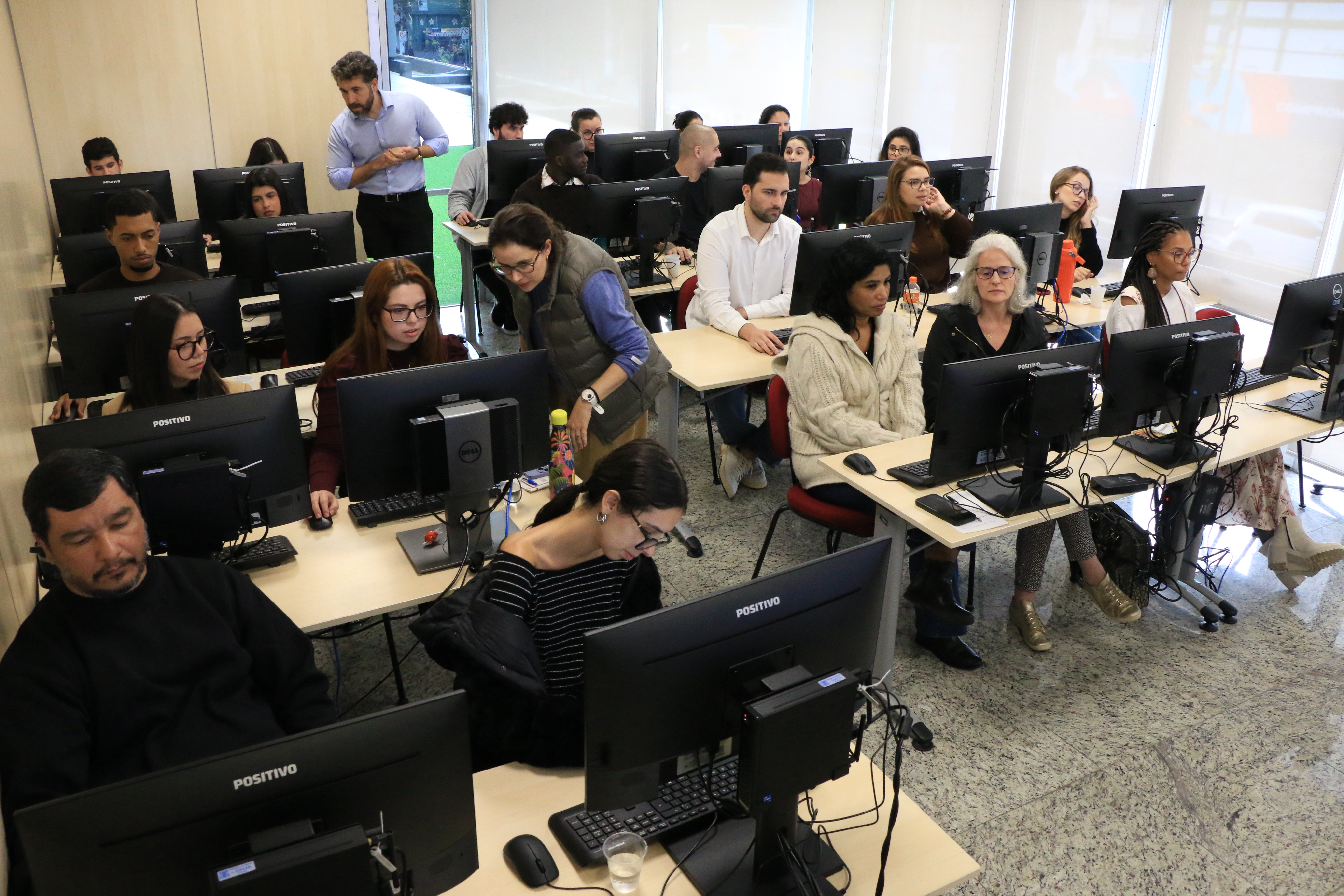 Foto horizontal da capacitação. A imagem mostra os participantes distribuídos em estações de trabalho equipadas com computadores, acompanhando atividades práticas relacionadas ao programa. Em destaque, os auditores fiscais de Controle Externo Michelle Padovese de Arruda e Jean Rodrigues de Souza orientam os participantes durante o treinamento, prestando esclarecimentos e auxiliando na execução das tarefas. 