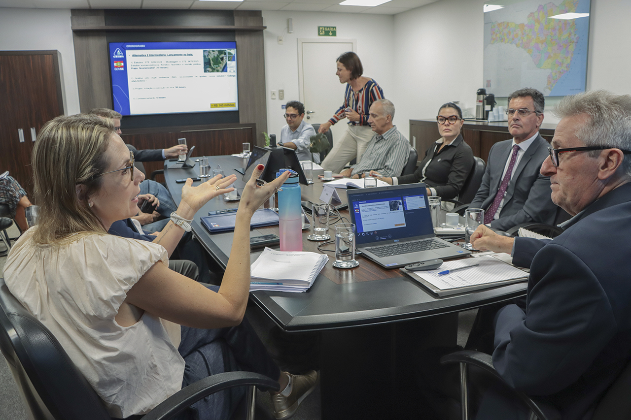 Foto da engenheira sanitarista da Casan, Andreia Senna Soares Trennepohl, falando aos demais durante a reunião, gesticulando. É uma mulher branca, loira e usa óculos de grau. Ao seu lado, outros participantes, entre eles, o coordenador da mesa, Neimar Paludo. 
