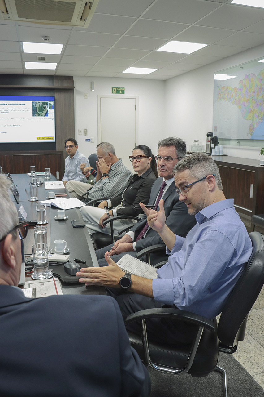 Foto do diretor da DEC, Maximiliano Mazera, falando aos demais durante a reunião. Ele é um homem branco, de cabelos grisalhos. Está ao lado do coordenador da mesa, Neimar Paludo, e do representante do MPSC, Luiz Fernando Ulysséa. 