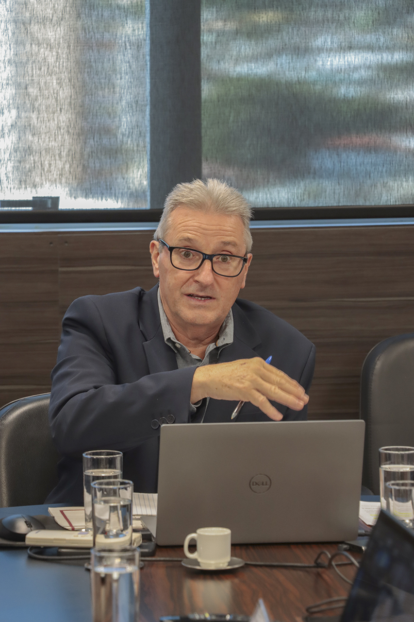 Foto do coordenador da mesa, Neimar Paludo, falando aos demais durante a reunião. Ele é um homem branco, de cabelos grisalhos. Veste paletó preto e usa óculos de grau.  