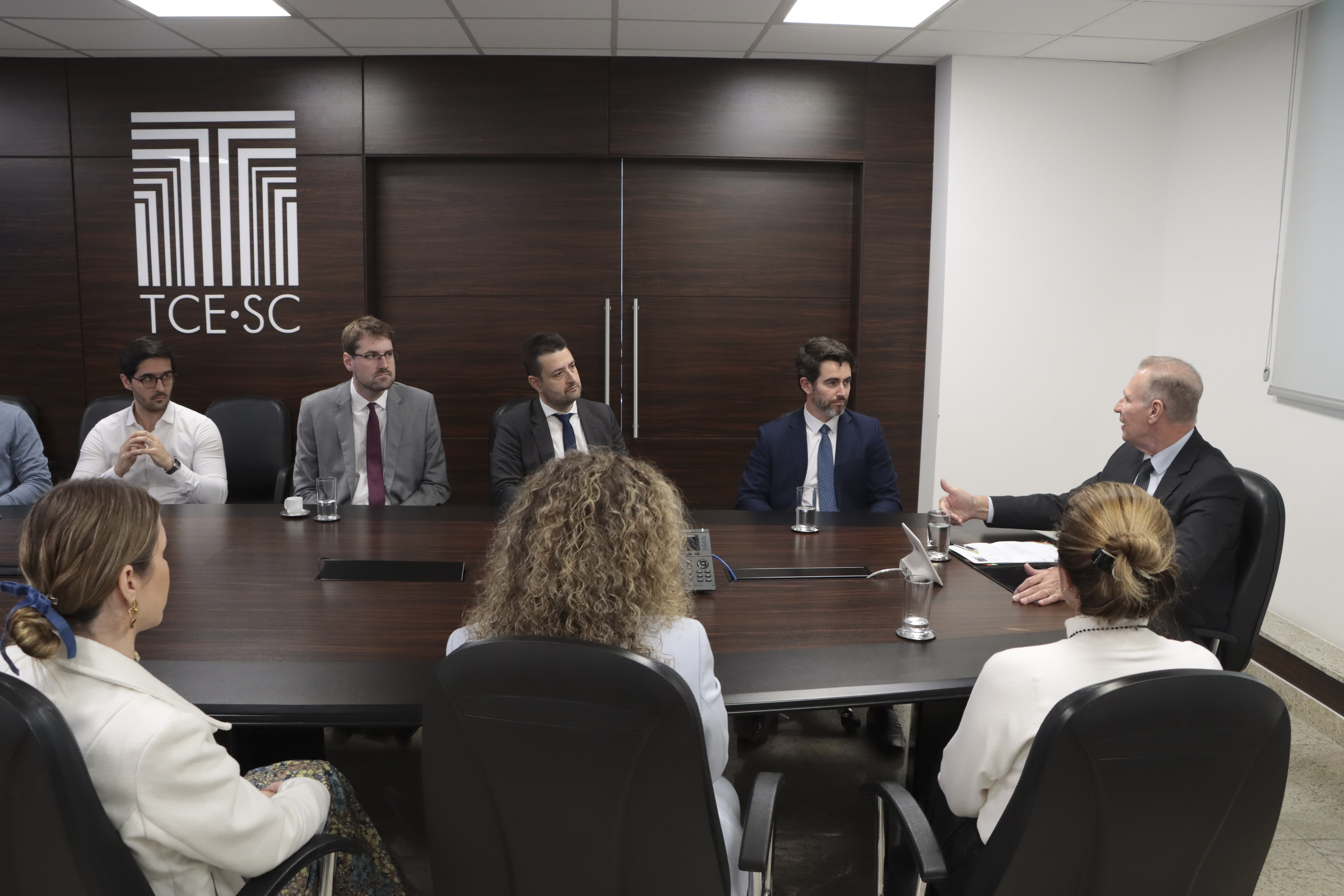 Foto horizontal que mostra vários participantes da reunião institucional, sentados lado a lado, diante da mesa. Há membros da PGE/SC e do TCE/SC. Na ponta, o conselheiro Herneus e o procurador Marcelo. Ao fundo, na parede, plotagem com logo do TCE/SC. 