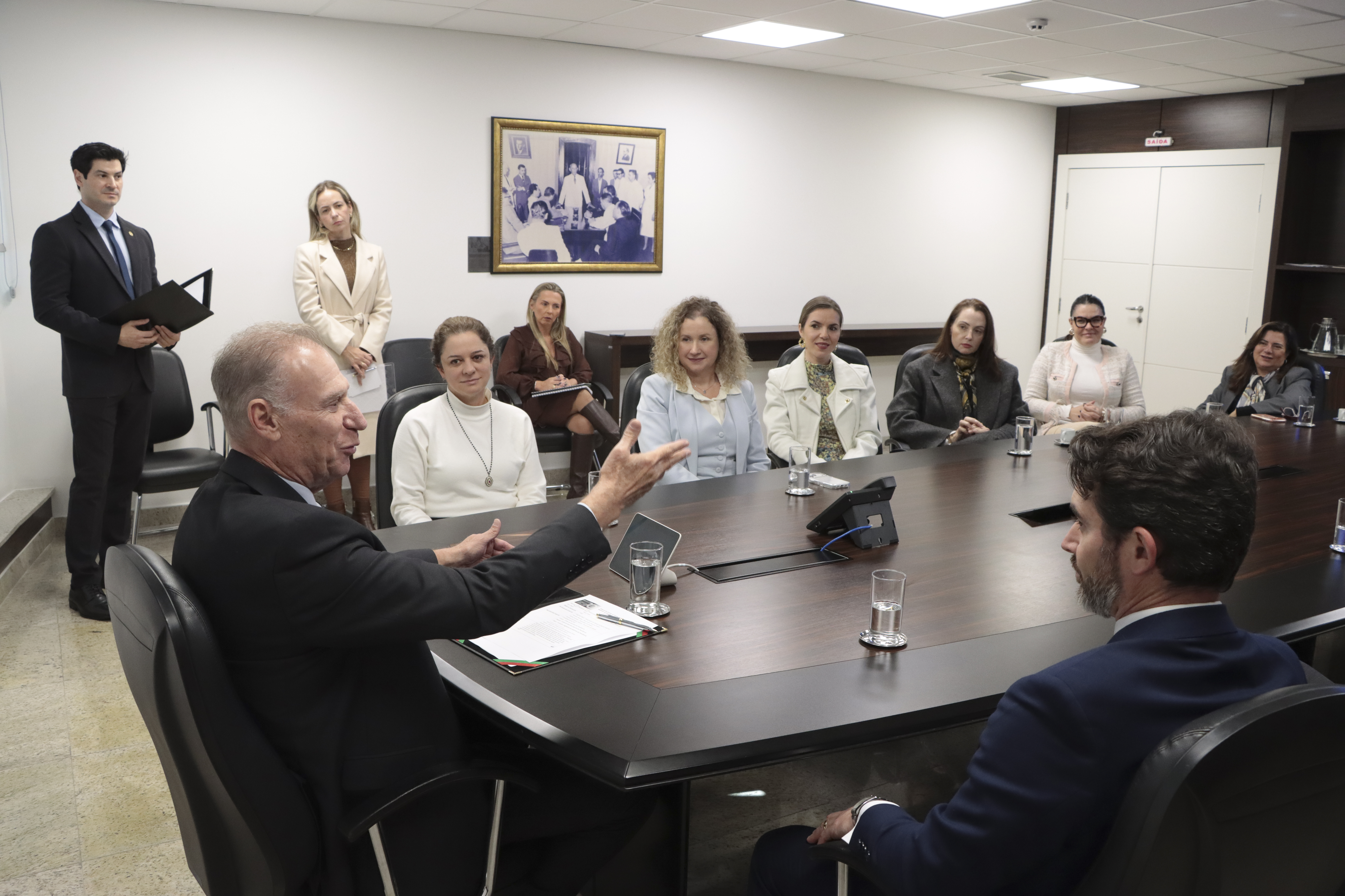 Foto horizontal dos demais participantes da reunião institucional. Em primeiro plano, o presidente Herneus e o procurador-geral Marcelo. Aparecem também, entre outros, a chefe de Gabinete da Presidência, Juliana Francisconi, a procuradora jurídica, Gláucia Mattjie e a secretária-geral, Flávia Baesso. Ao fundo, outros participantes observam a reunião. 