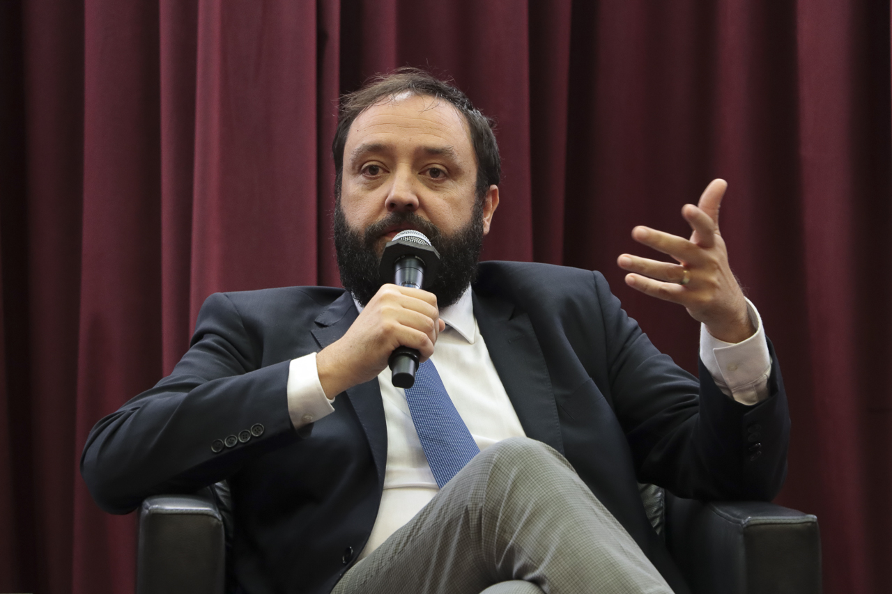 Foto horizontal do professor Caetano Dias. Ele é um homem branco, de cabelos e barba escuros. Veste terno e gravata e fala ao microfone, sentado em poltrona no palco do auditório do TCE/SC. Ao fundo, há cortinas bordôs. 