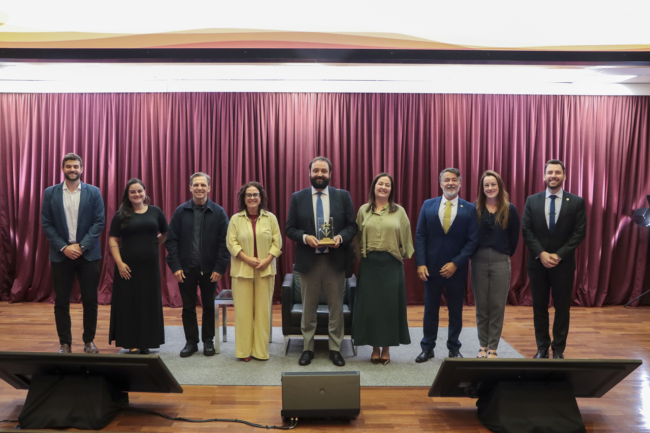 Foto horizontal dos organizadores do evento junto com o palestrante. Há, no palco do auditório, nove pessoas lado a lado. Entre eles, o professor Caetano, que segura um certificado recebido, e servidores do TCE/SC. 