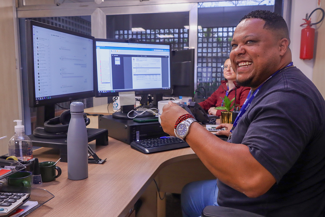 Foto do encarregado de almoxarifado Gustavo Fernando dos Santos, sentado diante de um computador, segurando sua caneca. Ele é um homem negro, de cabelos pretos. Veste uniforme azul e sorri para a foto. 