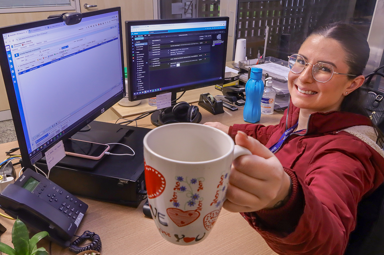 Foto da encarregada de almoxarifado Pamela Maestri Justino, segurando sua caneca. Ela é uma mulher branca, de cabelos pretos e presos. Ela está sentada diante de um computador, sorrindo. Veste roupa vermelha e levanta sua caneca, mostrando-a para a foto.  