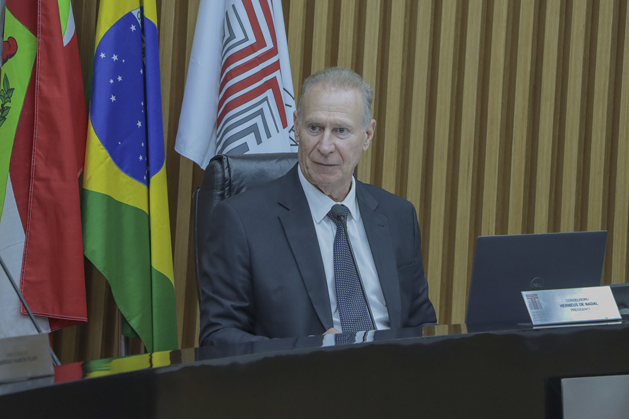 Banner horizontal com foto do presidente do TCE/SC, conselheiro Herneus De Nadal, durante sessão plenária. É um homem branco, de cabelos claros. Veste terno e gravata e está sentado diante da bancada de autoridades. Ao fundo, há bandeiras e um painel de madeira.  