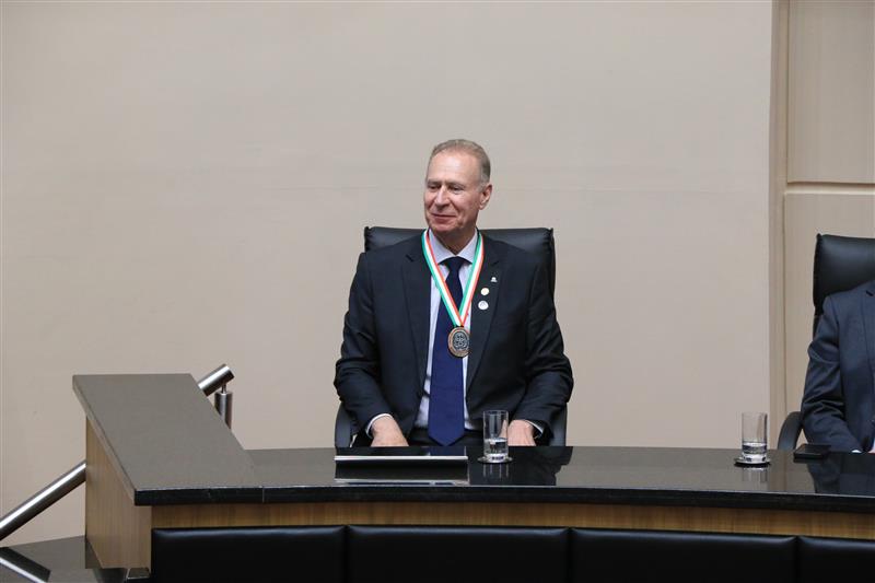 Foto do presidente do TCE/SC, conselheiro Herneus De Nadal, compondo a mesa de autoridades da sessão solene. Ele é um homem branco, de cabelos claros. Veste terno e gravata e usa a medalha recebida no pescoço.