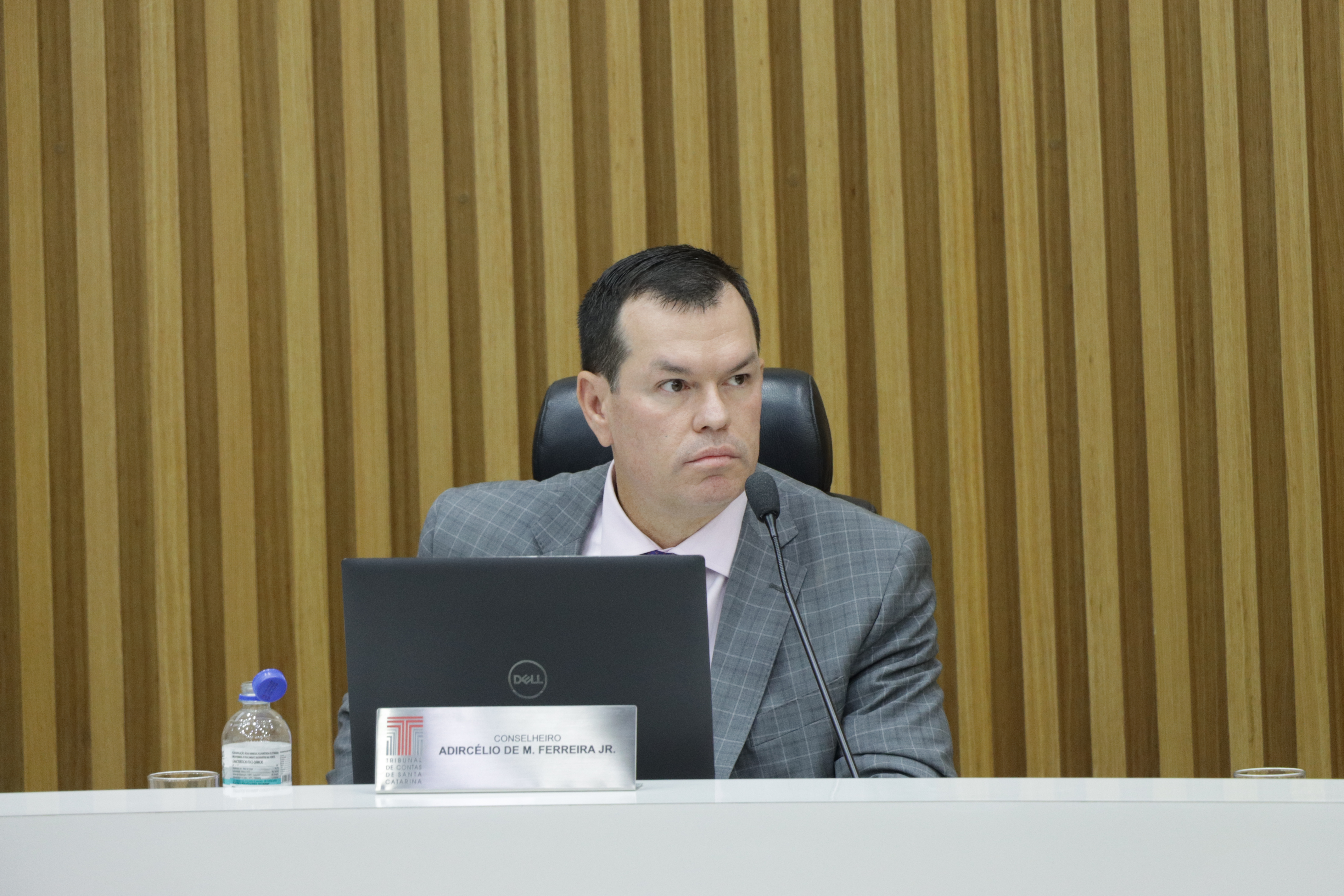Foto horizontal do conselheiro Adircélio Ferreira Júnior durante sessão plenária. É um homem branco, de cabelos escuros. Veste terno cinza e está sentado diante da bancada circular do Plenário, olhando para a direita. Ao fundo, painel amadeirado compõe o cenário.