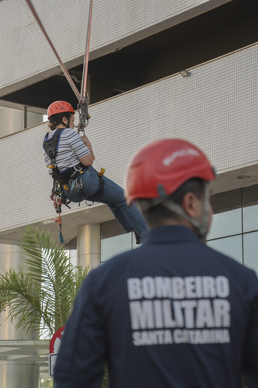 Foto vertical de uma pessoa descendo de tirolesa do prédio do TCE/SC, com todos os equipamentos de segurança. Em primeiro plano, um bombeiro militar de costas, que também usa capacete.