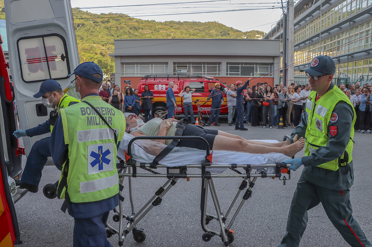 Foto horizontal de uma vítima interpretada por servidora do TCE/SC sendo transportada em maca do Corpo de Bombeiros para dentro da ambulância. Três bombeiros, que vestem uniforme neon, realizam o transporte.