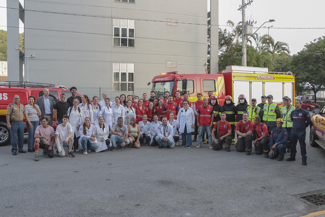 Foto horizontal que mostra a equipe que atuou no simulado de incêndio, lado a lado, na área externa do prédio do TCE/SC. Entre os presentes, bombeiros, os novos brigadistas, setor de saúde, vítimas interpretadas por servidores e Polícia Militar.