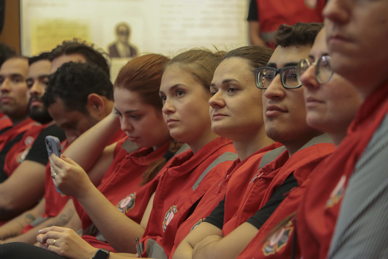 Foto horizontal que mostra parte dos novos brigadistas, lado a lado, sentados em poltronas do auditório. Eles vestem uniforme vermelho.