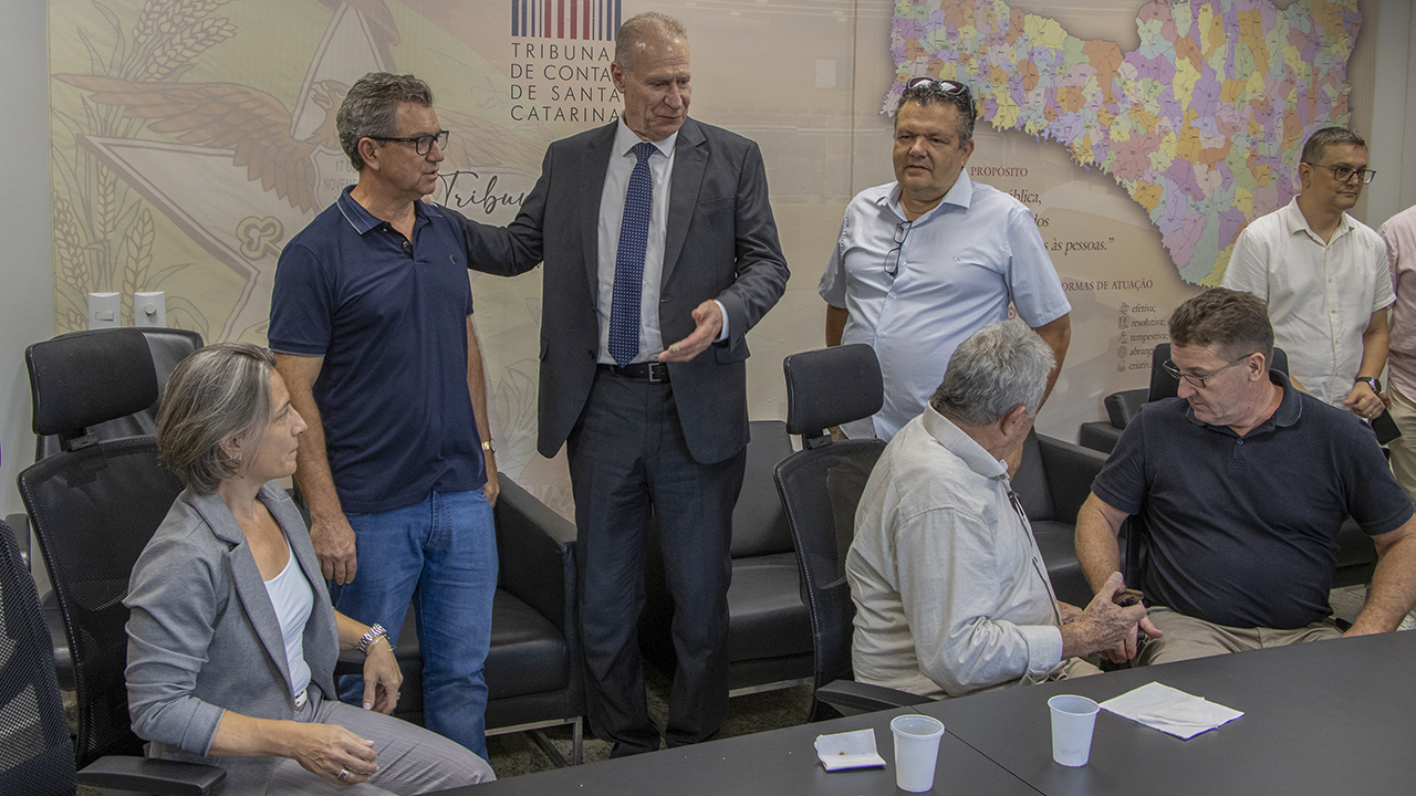 Imagem horizontal que mostra, ao centro, o presidente do TCE/SC, Herneus De Nadal, em pé, vestindo terno, enquanto conversa com os participantes. Ao seu lado, outros homens também estão em pé, e, ao redor da mesa, há pessoas sentadas. Ao fundo, aparece a identidade visual do TCE/SC e um mapa do estado de Santa Catarina. 