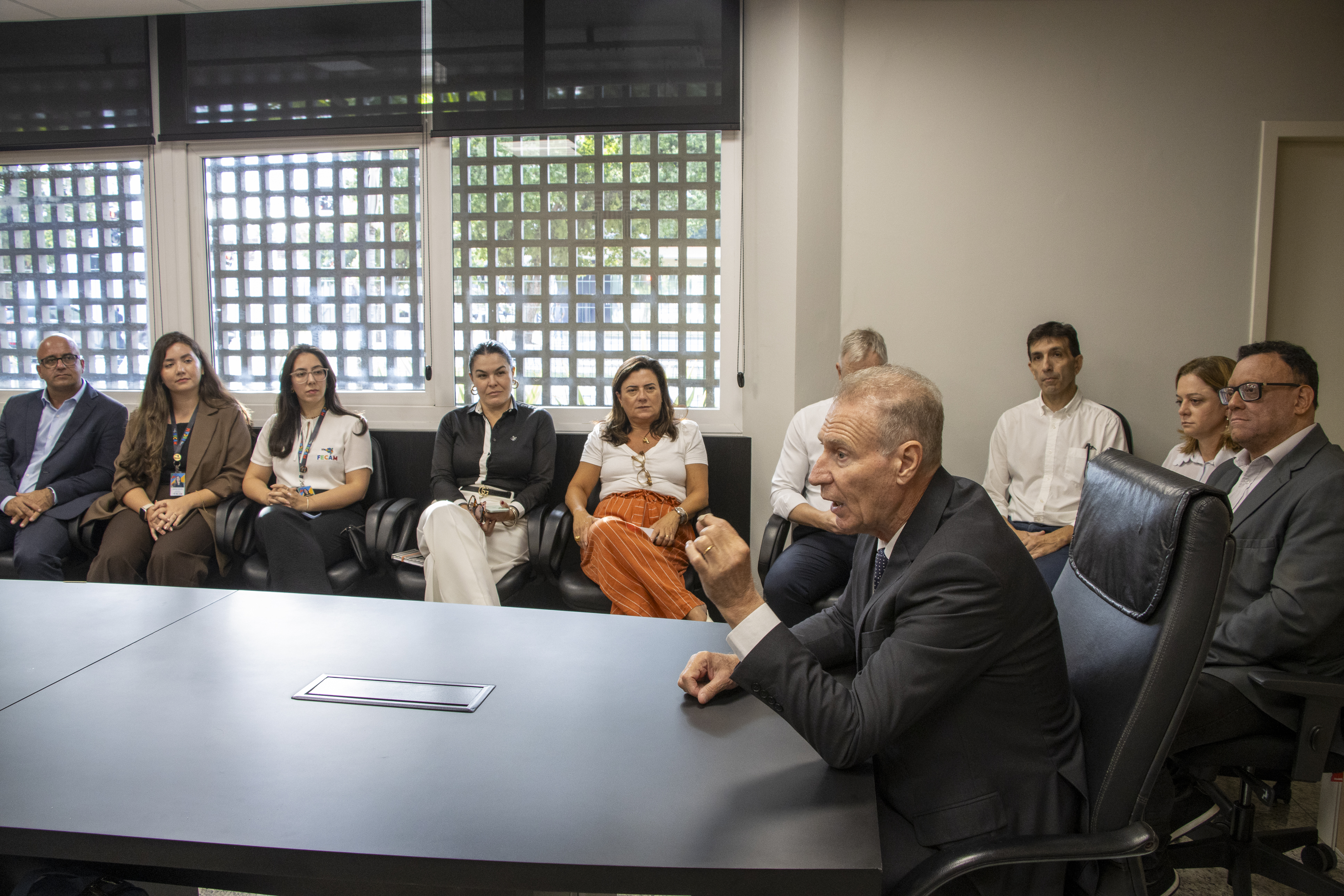 Foto horizontal que mostra uma reunião institucional em sala do TCE/SC, com participantes sentados ao redor de uma mesa retangular. Ao centro, o presidente Herneus De Nadal, em destaque, gesticulando enquanto fala. Ao redor, os demais acompanham a reunião com atenção. Na foto aparecem os diretores Monique Portella, Sidney Tavares Junior e Rogério Loch, além das chefes de gabinete Juliana Francisconi e Raquel Zomer. 
