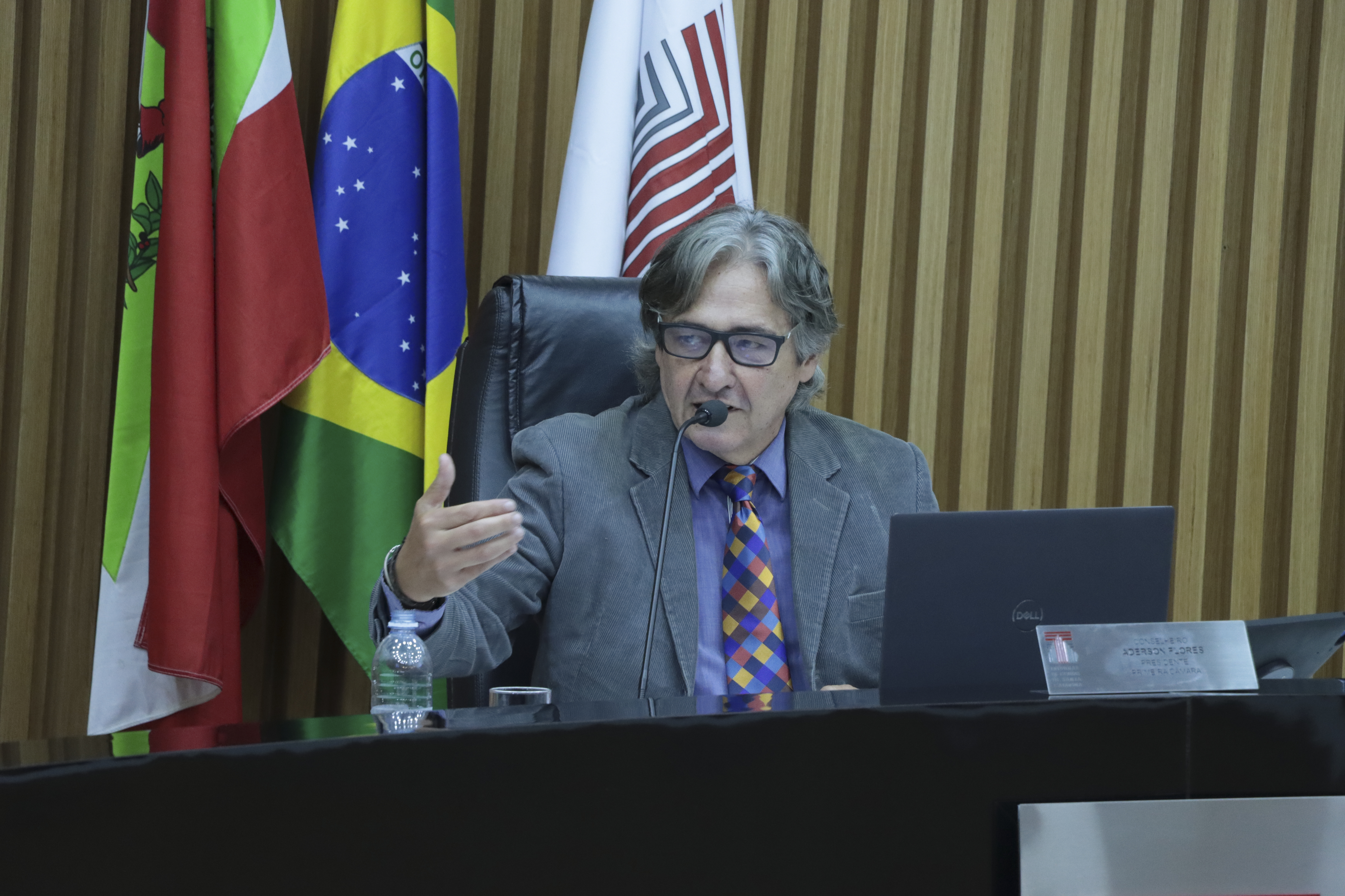 Imagem horizontal do presidente da Primeira Câmara, conselheiro Aderson Flores, durante sessão plenária. É um homem branco, de cabelos grisalhos. Veste terno cinza e fala ao microfone aos demais. Está sentado diante da bancada de autoridades. Ao fundo, há um painel de madeira e bandeiras do Brasil, de Santa Catarina e do TCE/SC. 