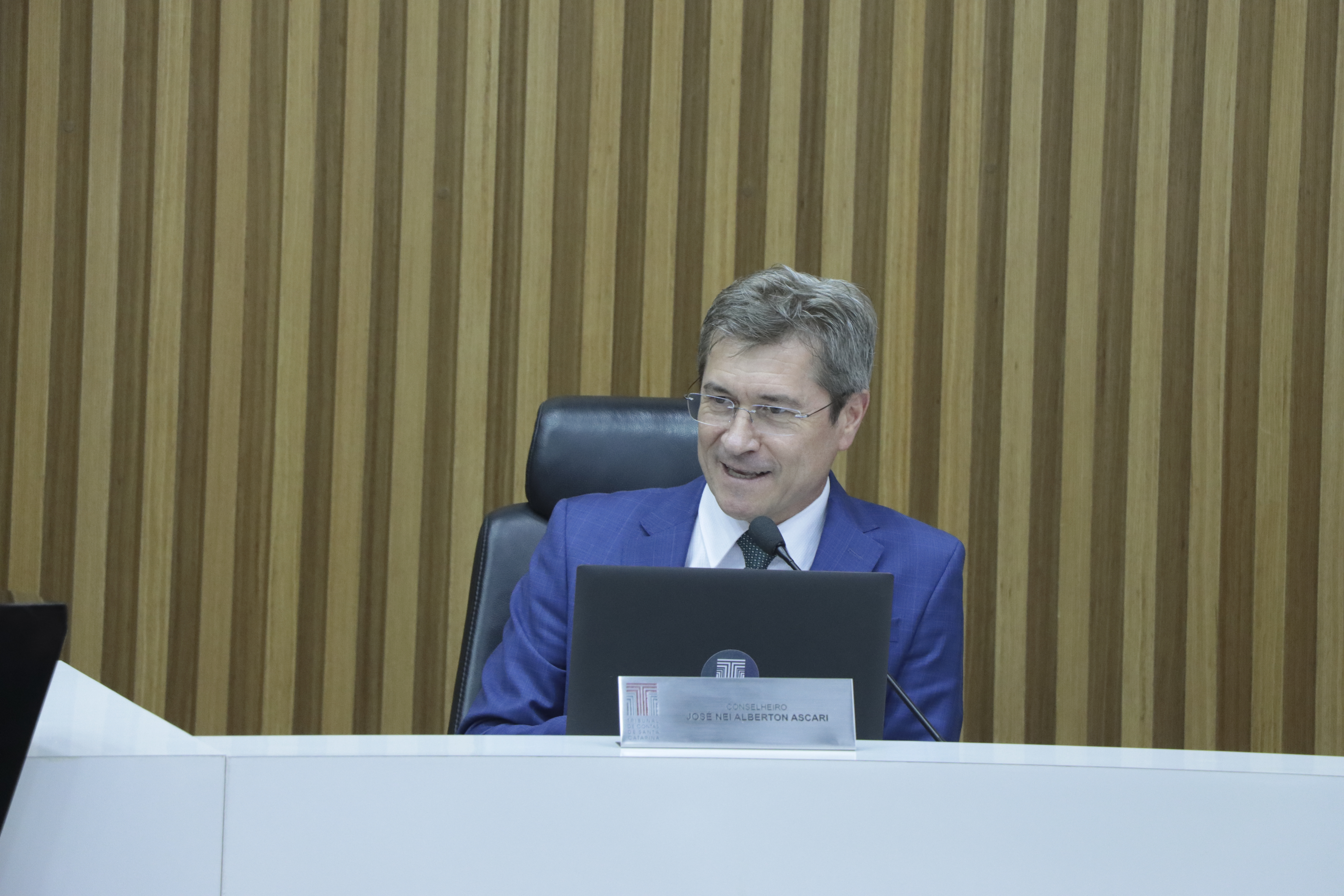 Imagem horizontal do conselheiro José Nei Ascari durante sessão plenária. É um homem branco, de cabelos claros. Veste terno azul e fala ao microfone aos demais. Está sentado diante da bancada de autoridades. Ao fundo, há um painel de madeira. 