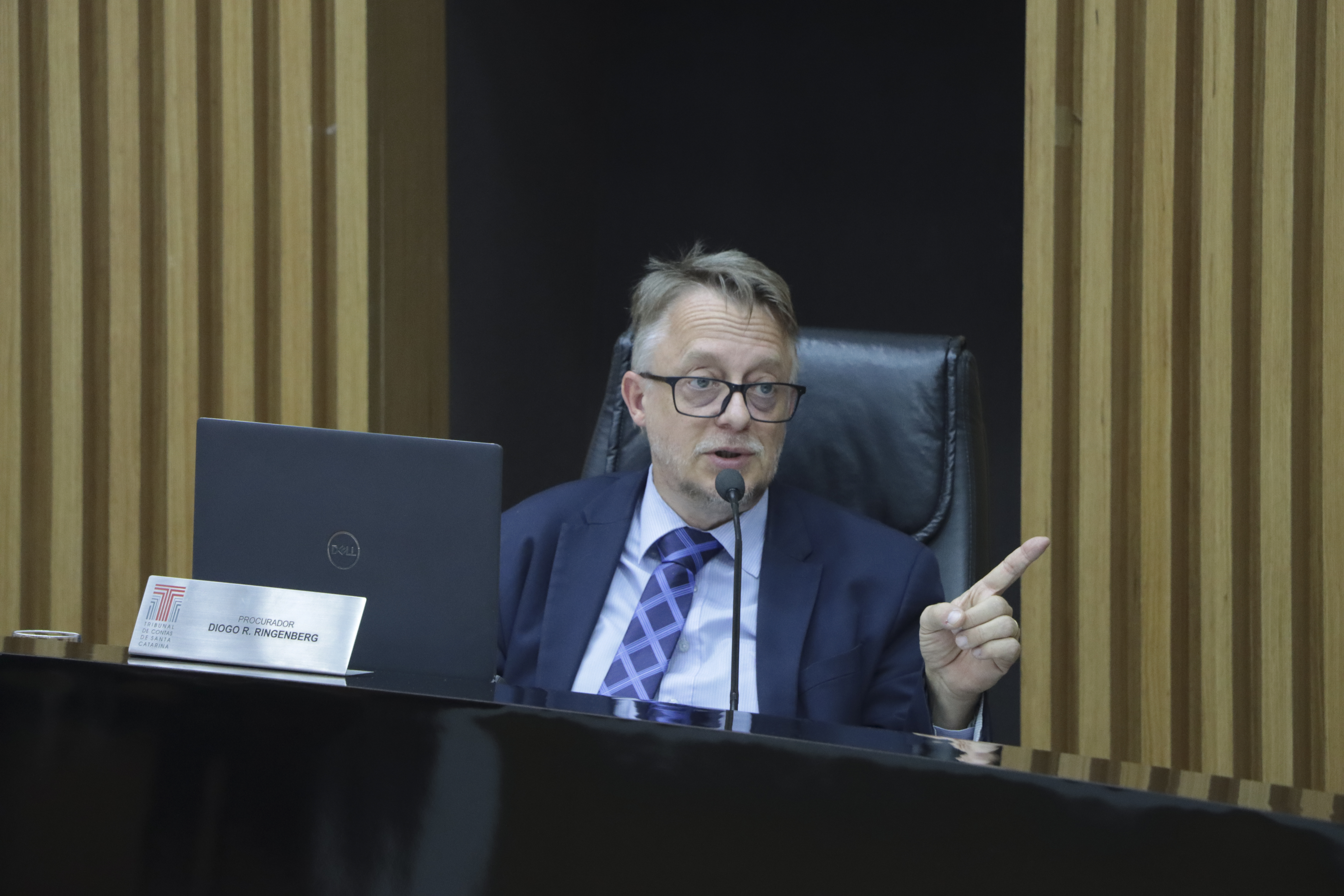 Imagem horizontal do procurador de Contas Diogo Ringenberg durante sessão plenária. É um homem branco, de cabelos claros. Veste terno preto e fala ao microfone aos demais. Está sentado diante da bancada de autoridades. Ao fundo, há um painel de madeira. 