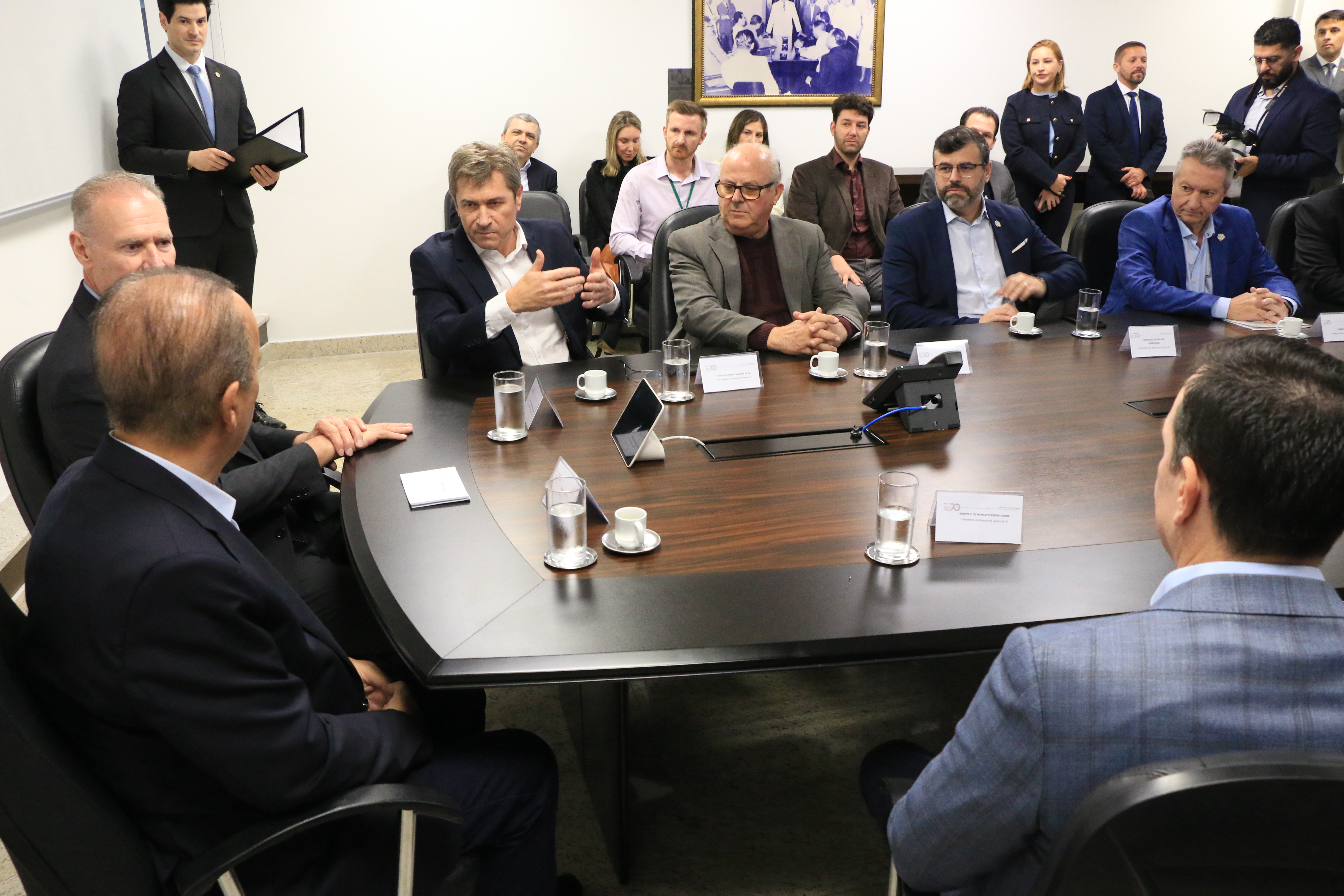 Foto horizontal que mostra vários participantes sentados em volta da mesa durante a reunião institucional. Entre os presentes, o presidente Herneus, o governador Jorginho e o conselheiro Ascari, que fala aos demais. Ao fundo, outras pessoas acompanham e registram a reunião. 