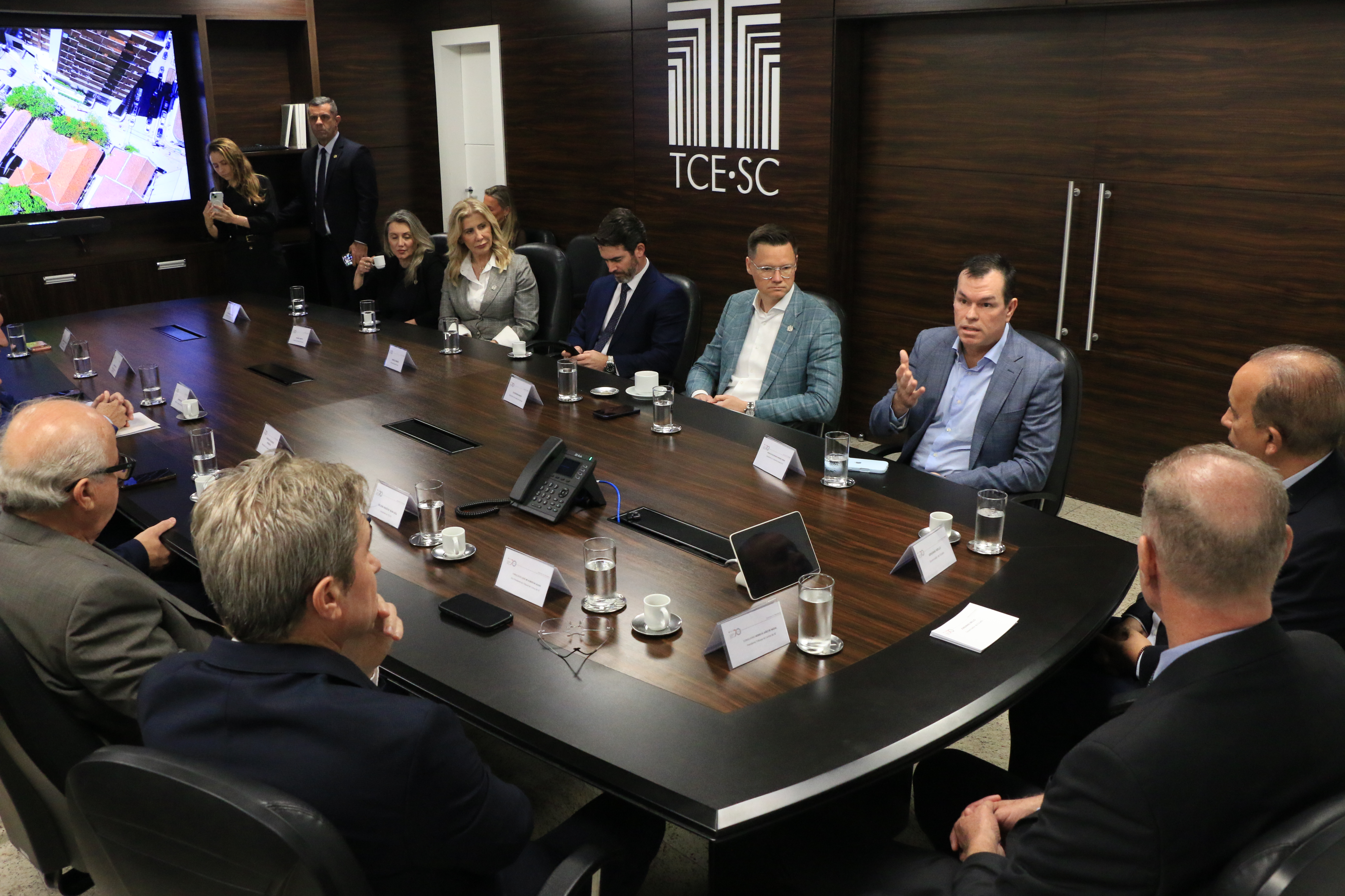 Foto horizontal que mostra parte dos participantes da reunião realizada. Entre os presentes, o conselheiro Adircélio, que fala aos demais, e os secretários Cleverson Siewert e Luciane Ceretta. Eles estão lado a lado, sentados diante da mesa.