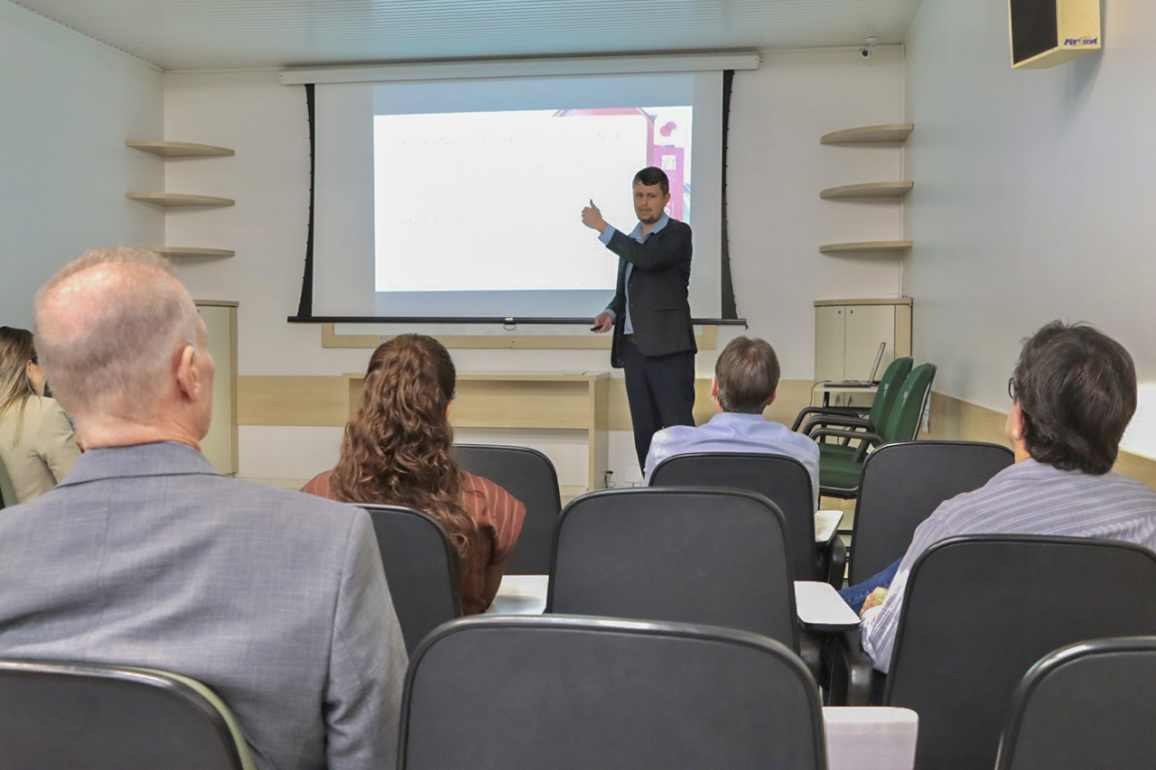 Foto horizontal que mostra o auditor fiscal de Controle Externo Gabriel Schiochet durante apresentação em Chapecó, posicionado à frente de uma sala de treinamento, gesticulando enquanto se dirige ao público. À frente, há participantes sentados em cadeiras organizadas em fileiras, voltados para a apresentação projetada em tela, compondo o ambiente institucional de capacitação. 