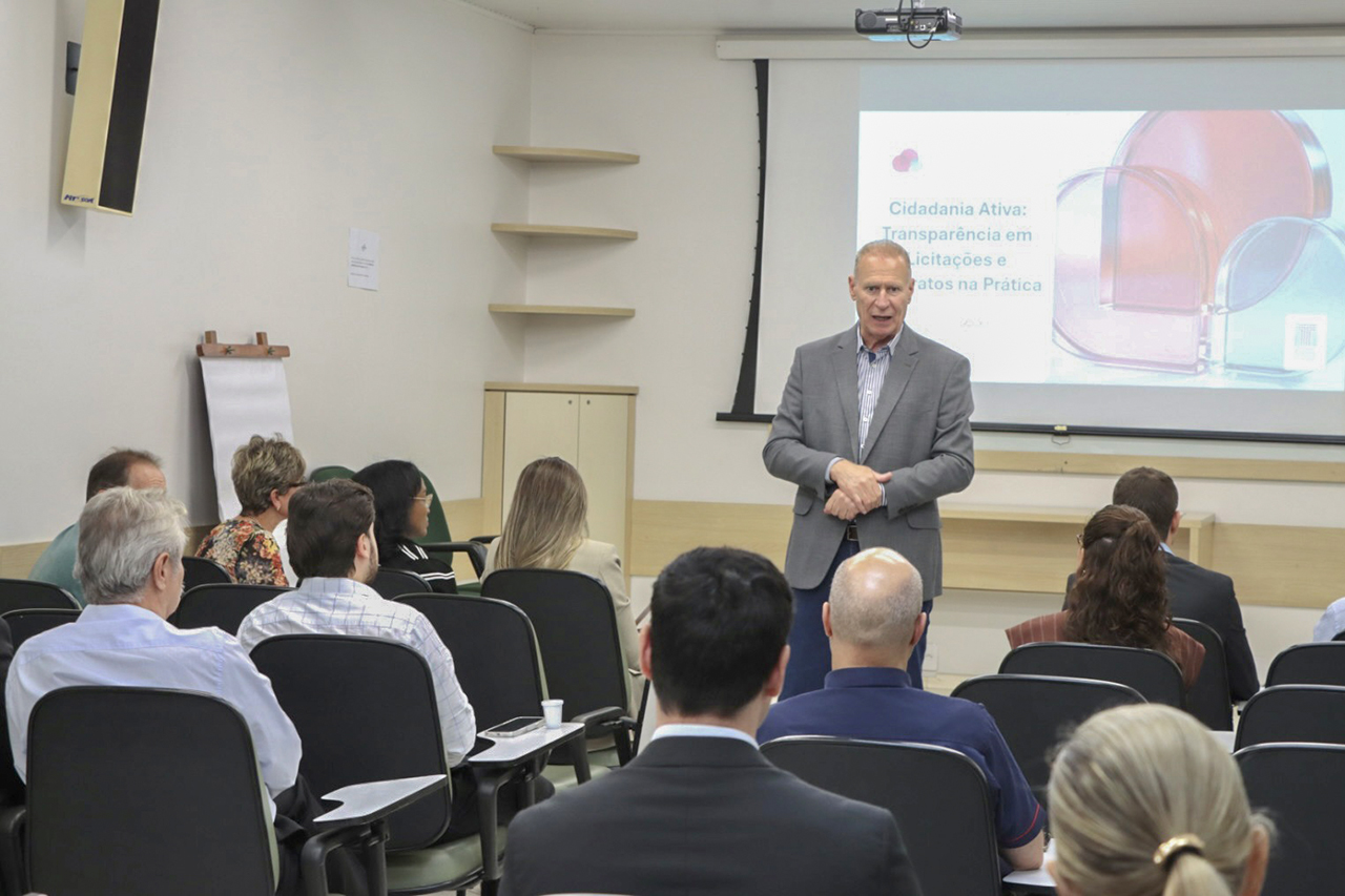 Foto horizontal que mostra o presidente Herneus De Nadal durante fala em capacitação em Chapecó, posicionado à frente de uma sala de treinamento e se dirigindo ao público. À frente, participantes estão sentados em cadeiras organizadas em fileiras, voltados para a apresentação projetada em tela ao fundo, que exibe conteúdo institucional, compondo o ambiente de aprendizado e interação. 