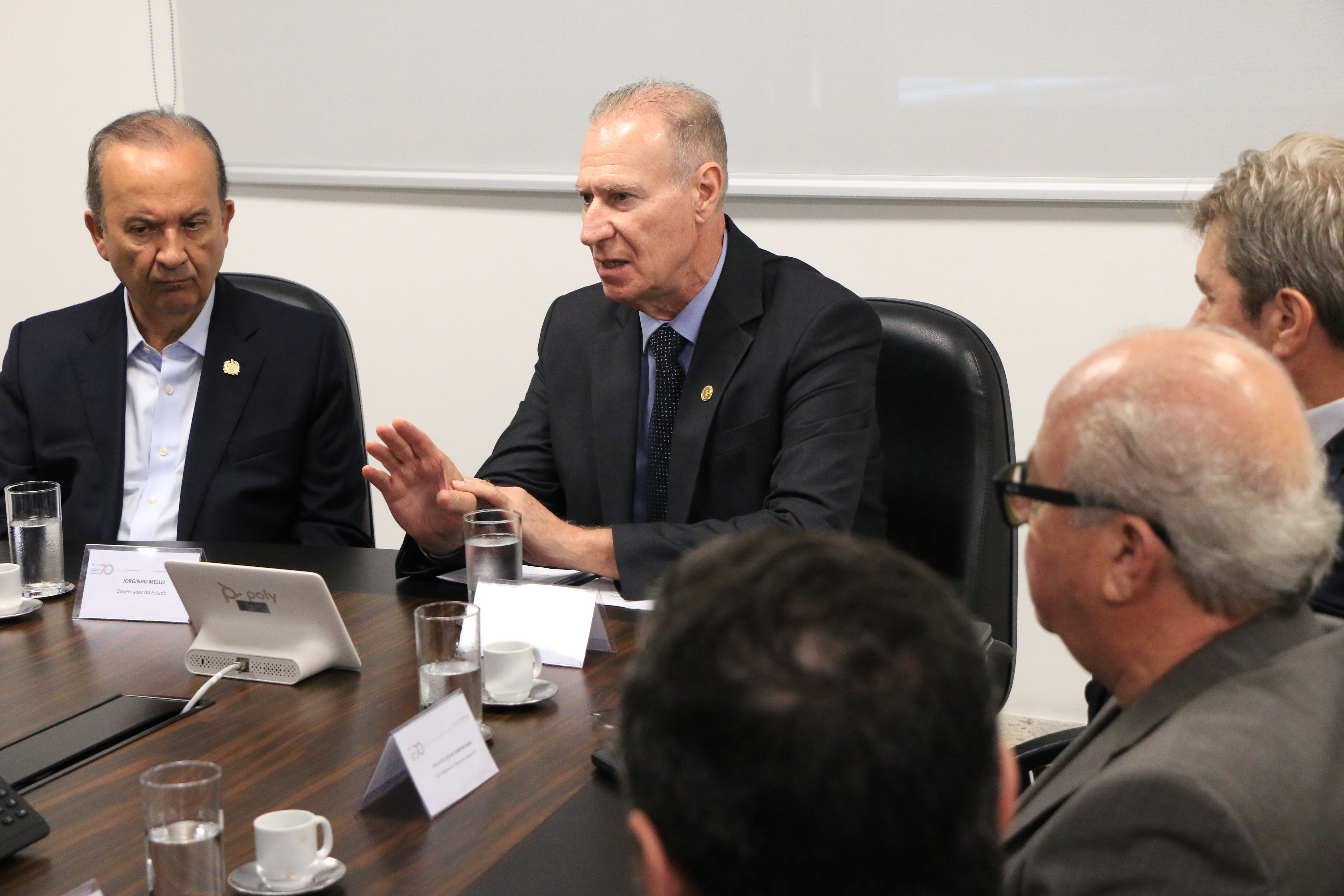 Foto horizontal do presidente do TCE/SC, conselheiro Herneus De Nadal, ao lado do governador Jorginho Mello, falando aos demais em reunião. Há várias pessoas sentadas diante de uma mesa no TCE/SC, enquanto conversam. 