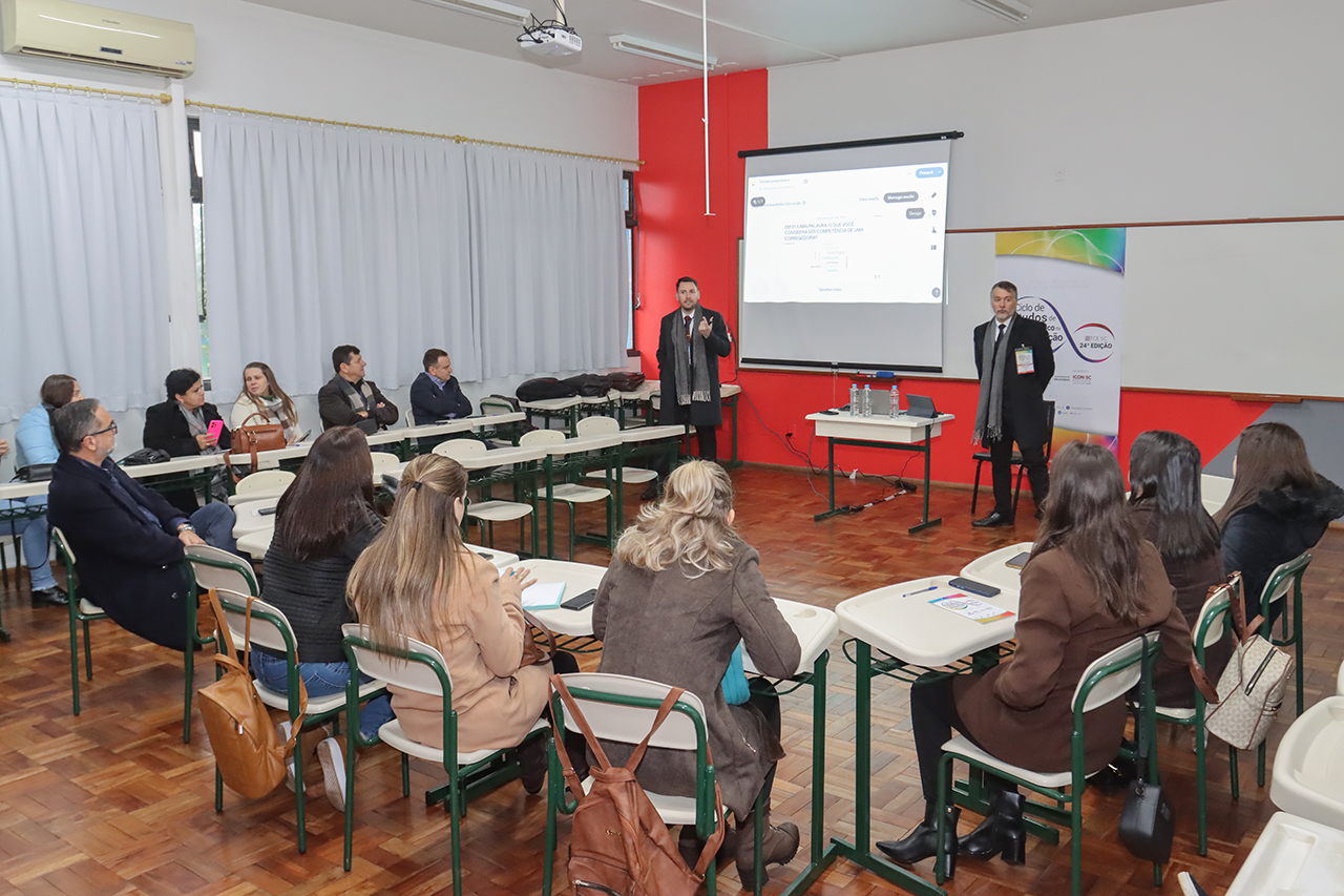 Foto de uma das oficinas do Ciclo de Estudos. Há vários participantes, sentados em cadeiras que formam um semicírculo em uma sala de aula. À frente deles, os servidores do TCE/SC Rogério Guilherme de Oliveira e Geovane Eziel Cardoso, que estão em pé, coordenando as atividades. Ao fundo, projeção de apresentação.