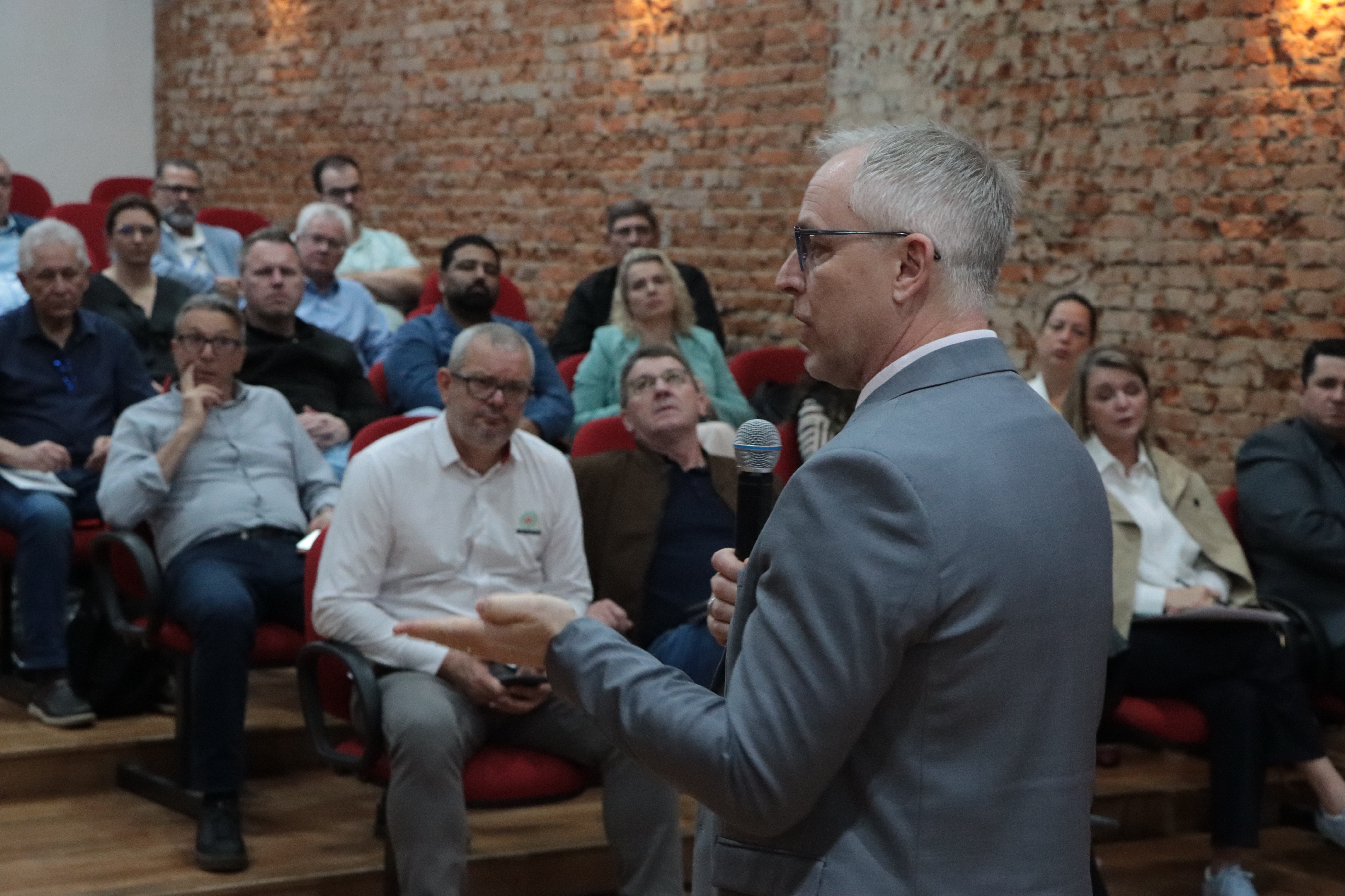 Banner horizontal com foto, de perfil, do diretor Sidney Tavares. Ele é um homem branco, de cabelos grisalhos. Veste paletó cinza e fala ao microfone, no palco. Ao fundo, acompanham a apresentação os participantes do evento, sentados lado a lado em poltronas.