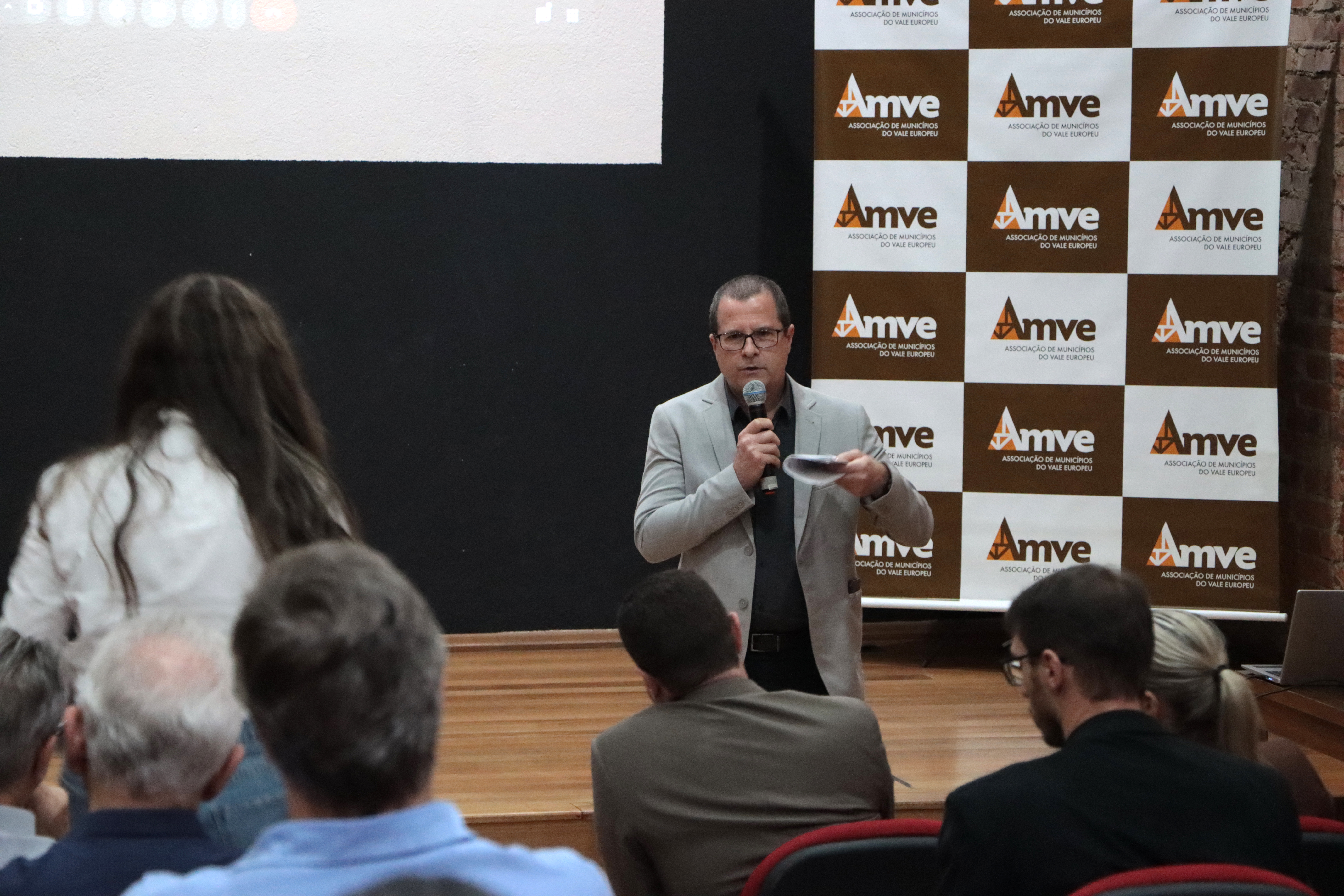 Foto do diretor Nilsom falando ao público, em pé, no palco do auditório. Atrás dele, banner com logo da Amve. À sua frente, público de costas, assistindo à apresentação.