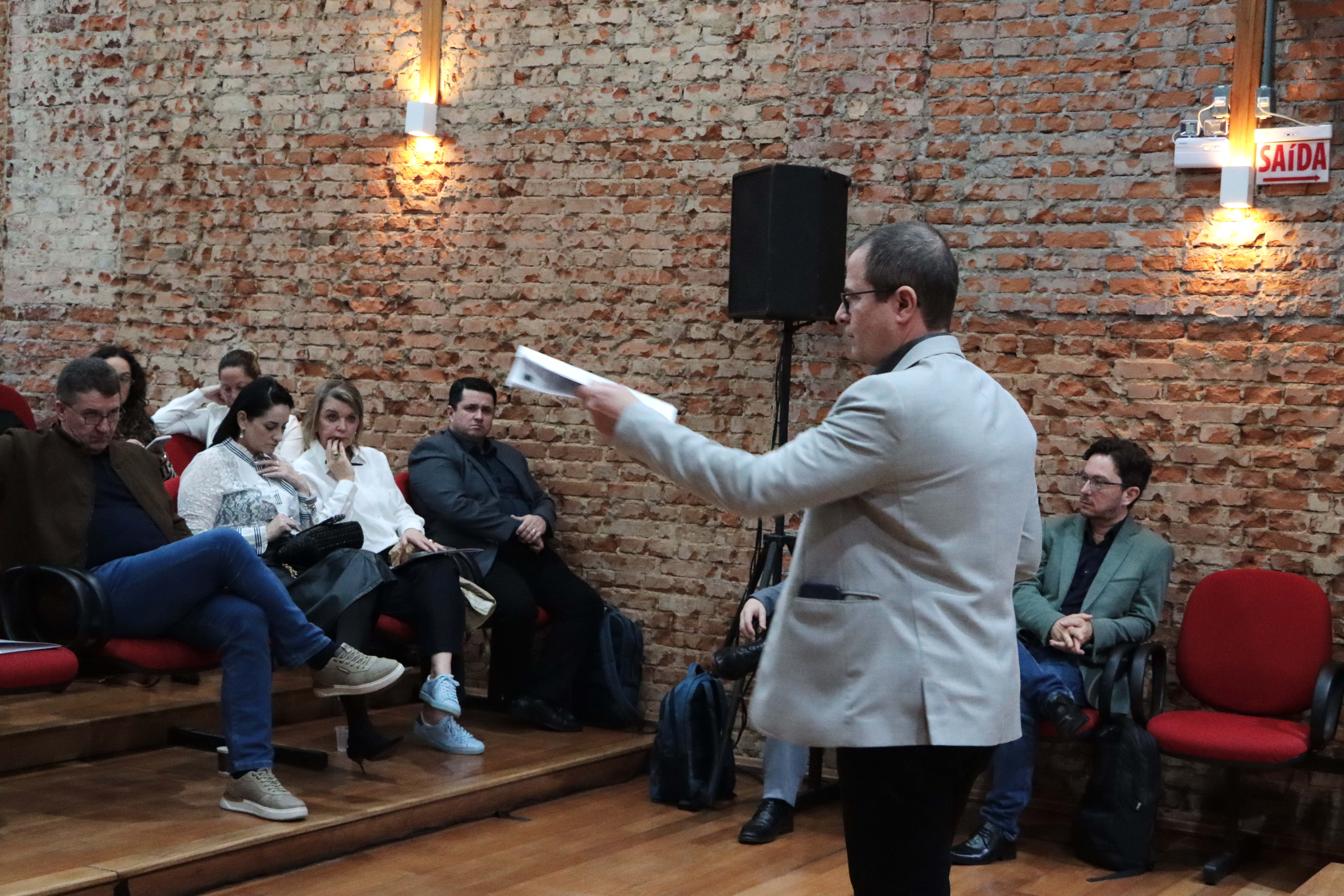 Foto horizontal do diretor Nilsom falando ao público durante o evento. É um homem branco, de cabelos escuros. Veste paletó claro. Está em pé, de costas para a foto. À sua frente, o público acompanha a palestra.