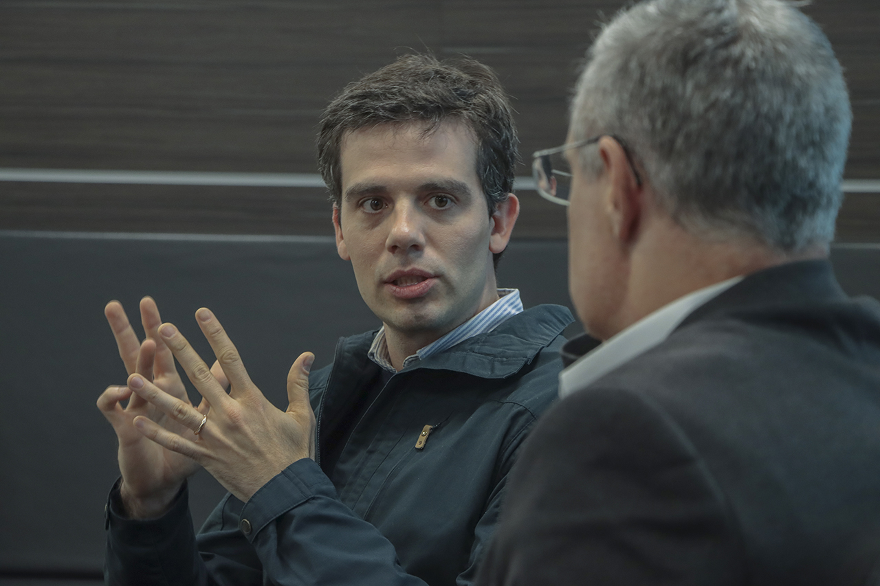 Foto horizontal do representante do Todos Pela Educação, Gabriel Barreto, falando aos demais. É um homem branco, de cabelos escuros. Gesticula enquanto fala. Ao seu lado, de costas para a foto, o conselheiro substituto Gerson Sicca. 