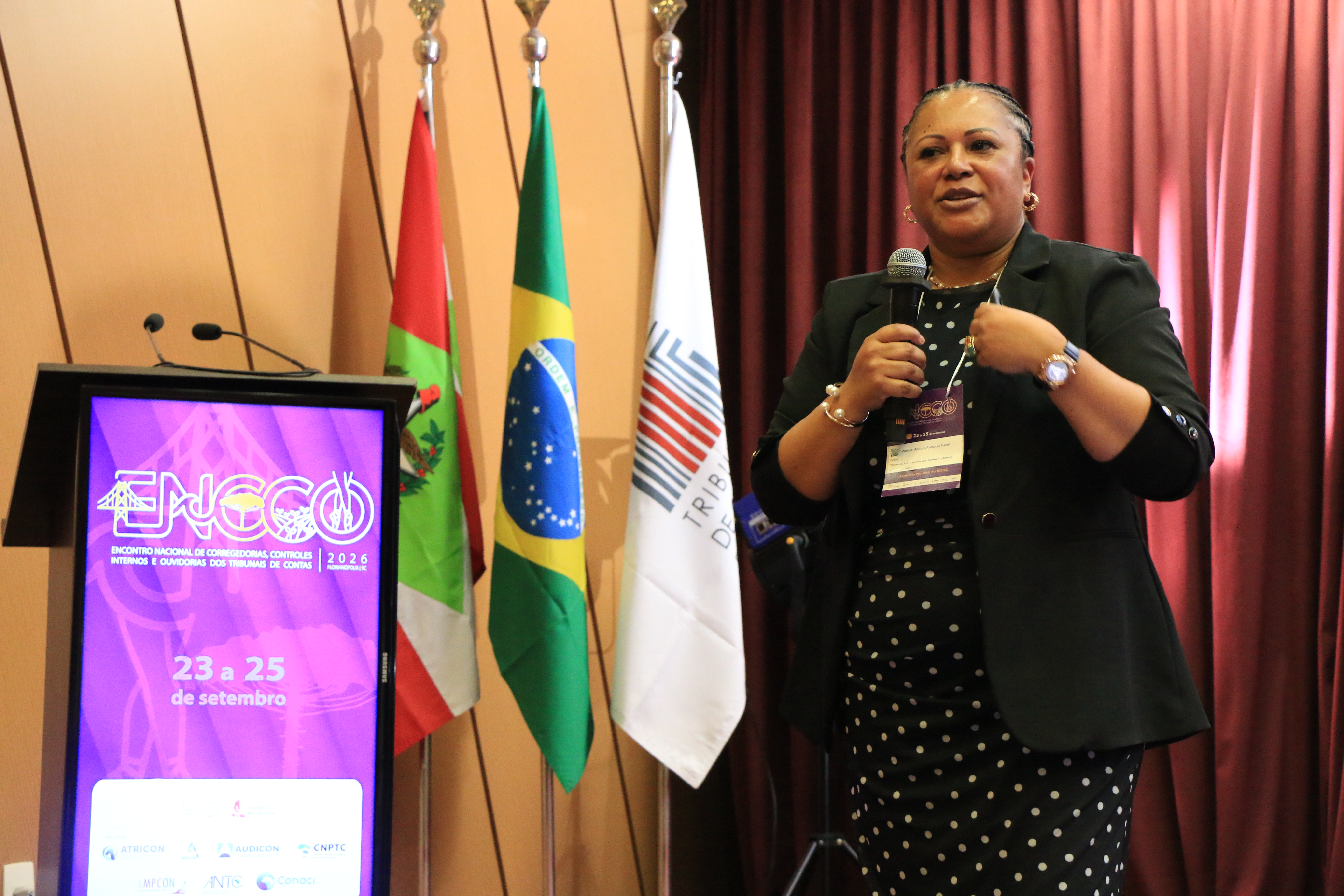 Foto horizontal da diretora da Ouvidoria, Walkíria Maciel, falando ao microfone durante o evento. Ela é uma mulher negra, de cabelos escuros, que estão presos. Veste roupa preta e está em pé, próxima ao púlpito.