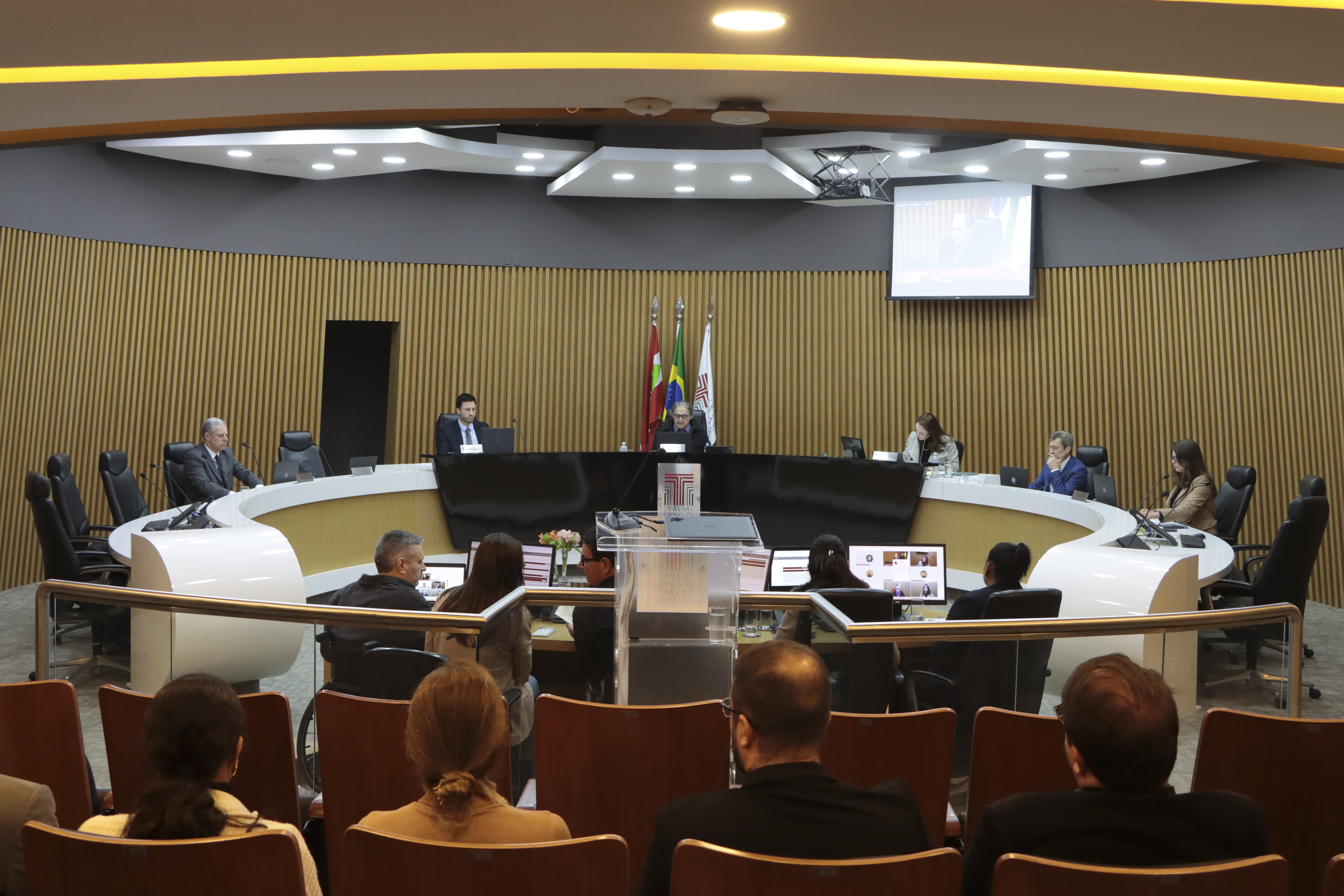 Foto horizontal dos membros da Primeira Câmara durante sessão no Plenário do TCE/SC. O ambiente tem uma bancada circular, diante da qual estão posicionados os conselheiros. Ao centro, o presidente da Primeira Câmara, Aderson Flores, guia os trabalhos. Em primeiro plano, de costas, alguns participantes na plateia. 